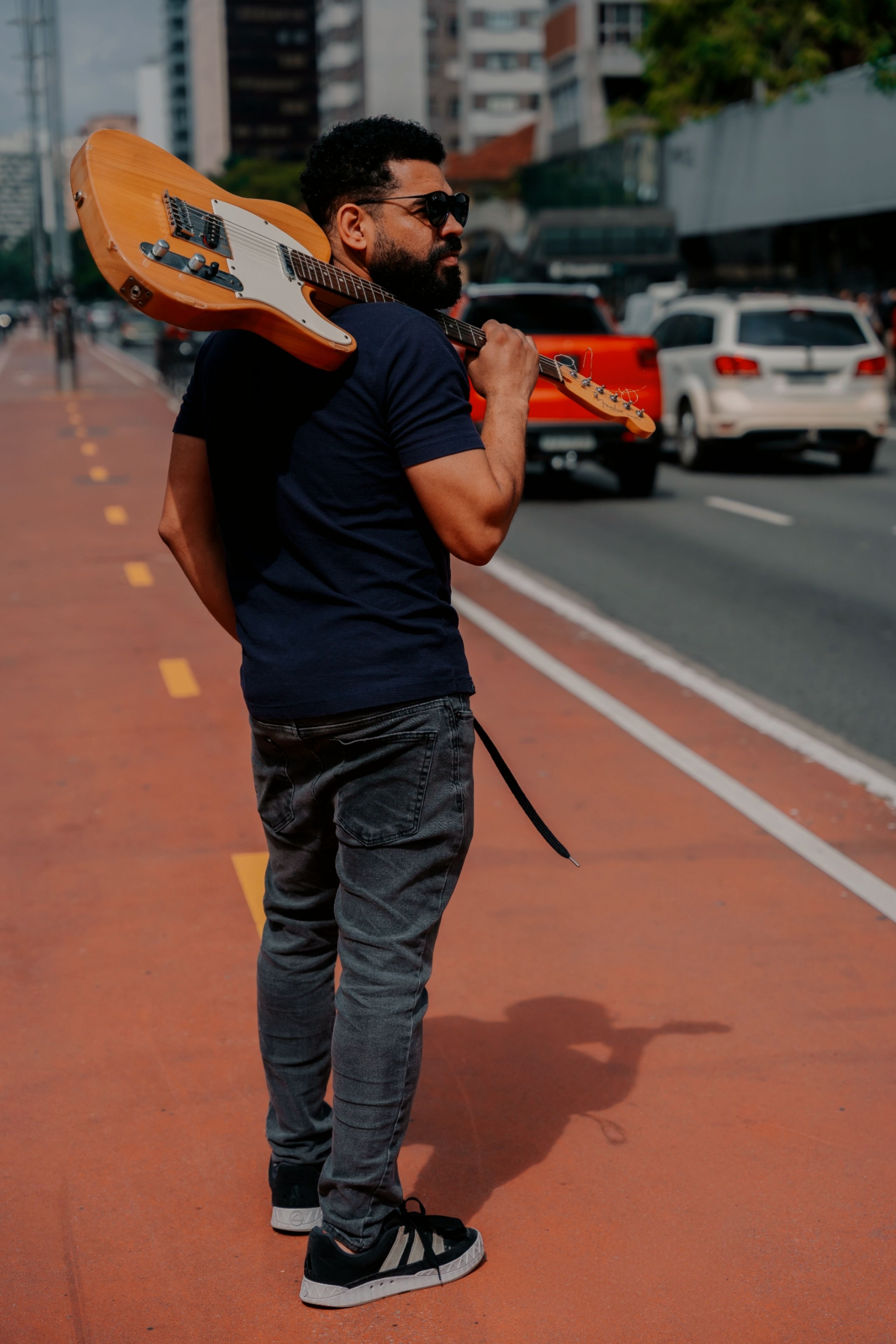 Young man carrying guitar on shoulder walking down a lively city street.