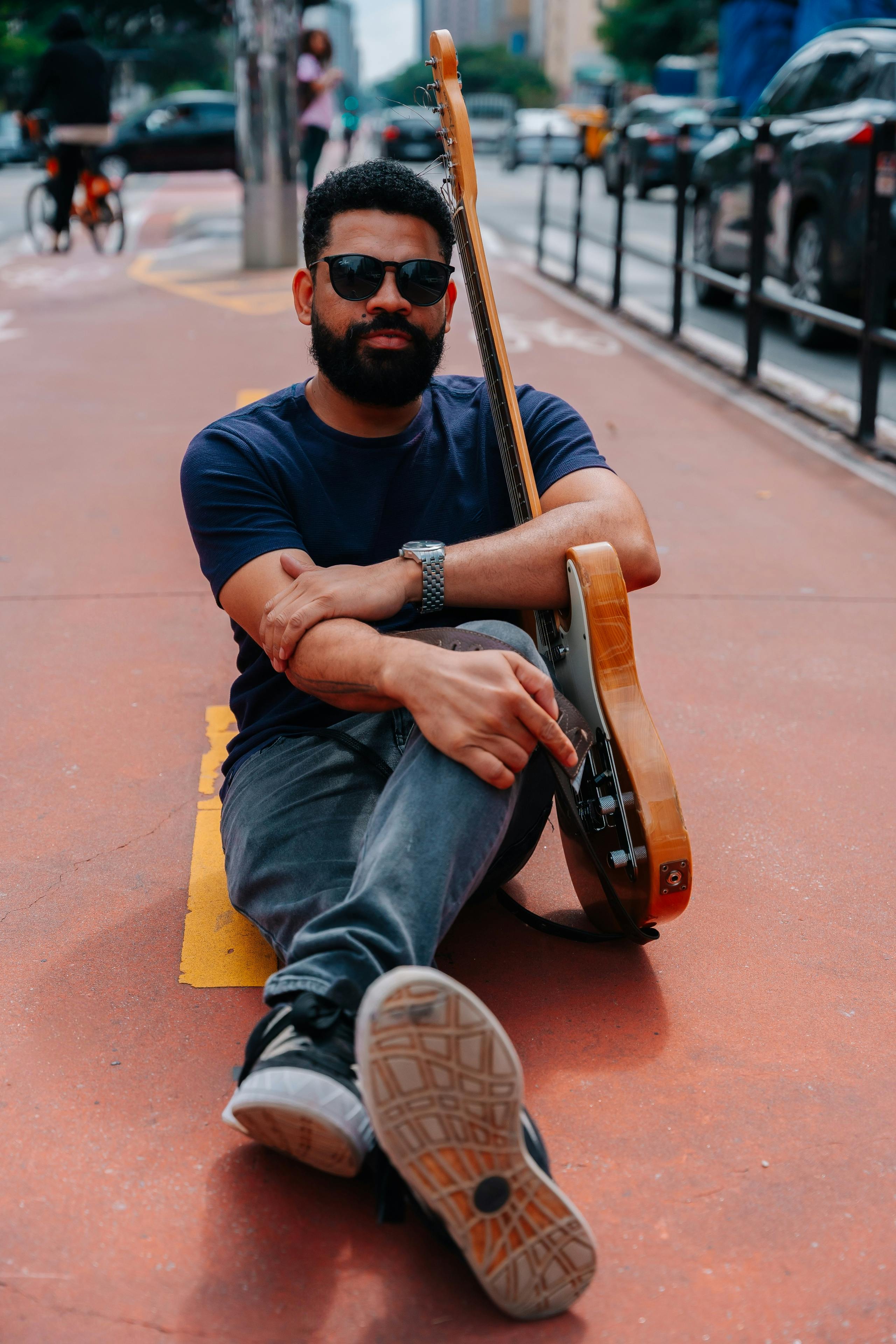Man sitting with guitar on city street, casual style, sunglasses.