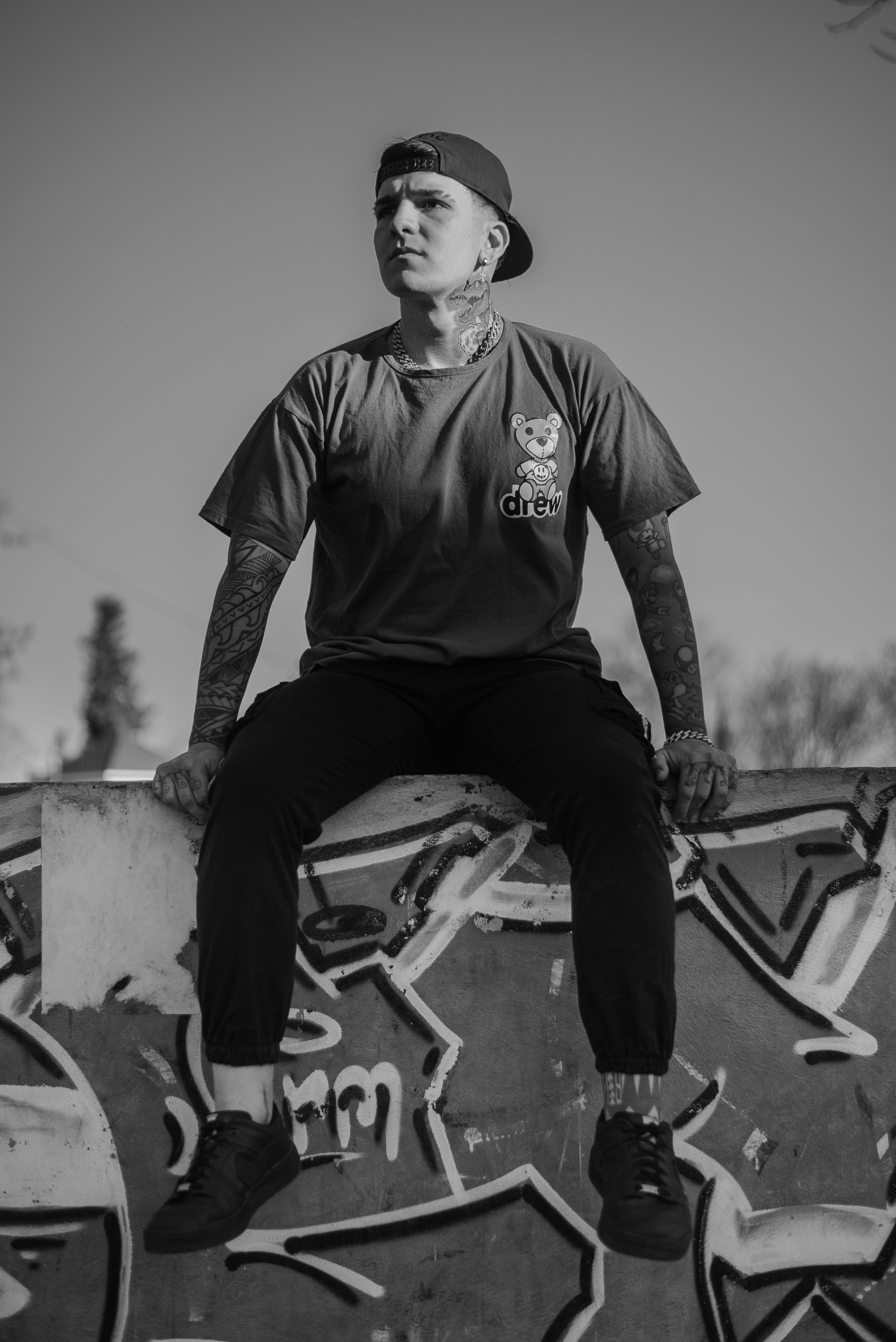 Monochrome photo of a tattooed individual sitting on a graffiti wall, exuding urban style.
