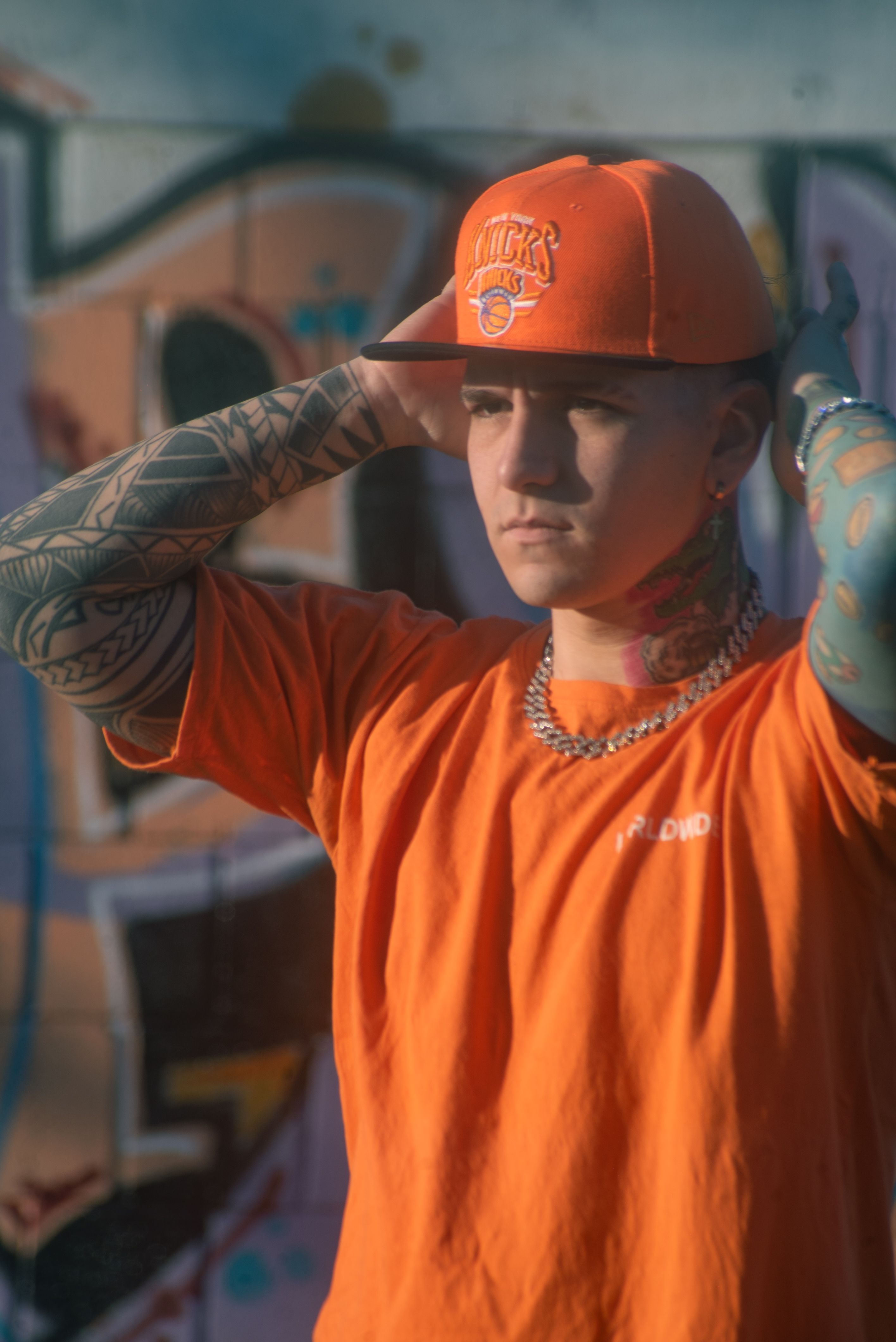 A young man wearing orange streetwear stands confidently in front of a colorful graffiti wall, embodying urban culture.