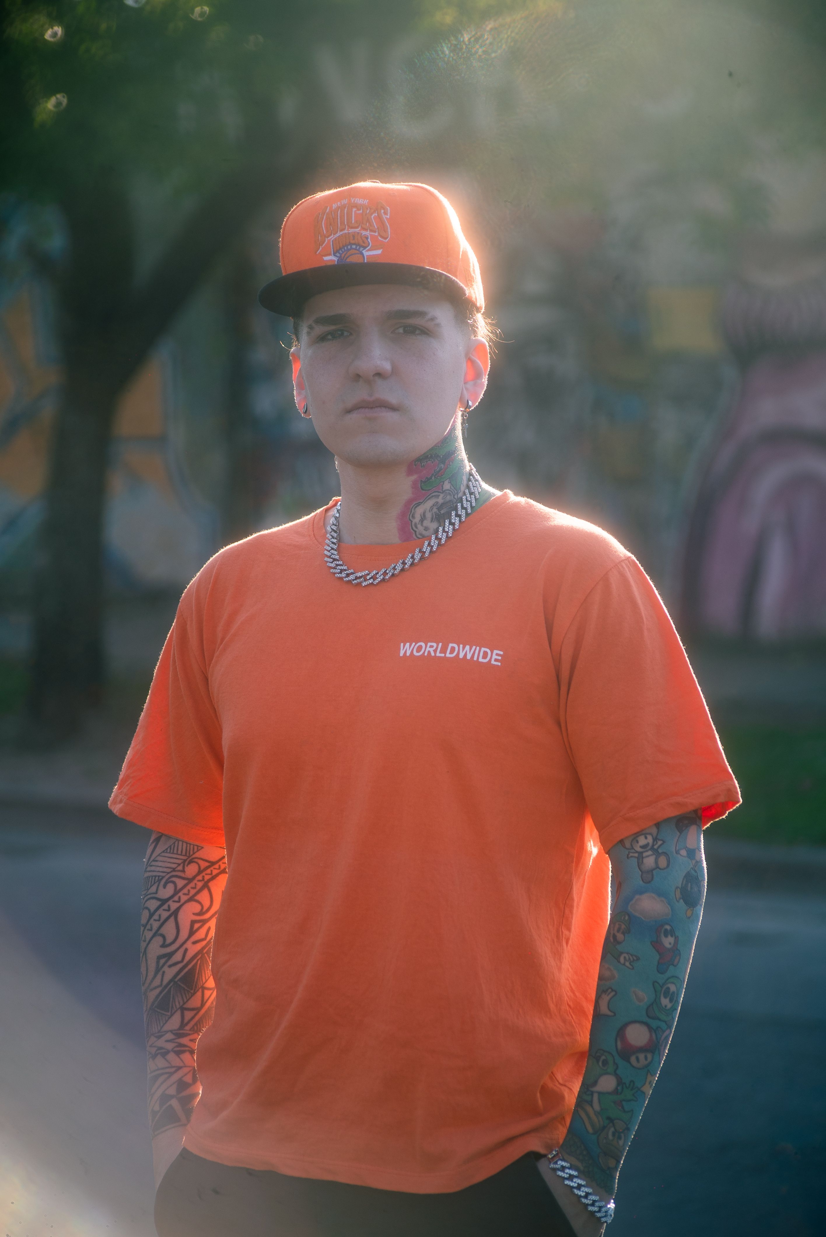 A young man with tattoos stands confidently outdoors wearing an orange shirt.