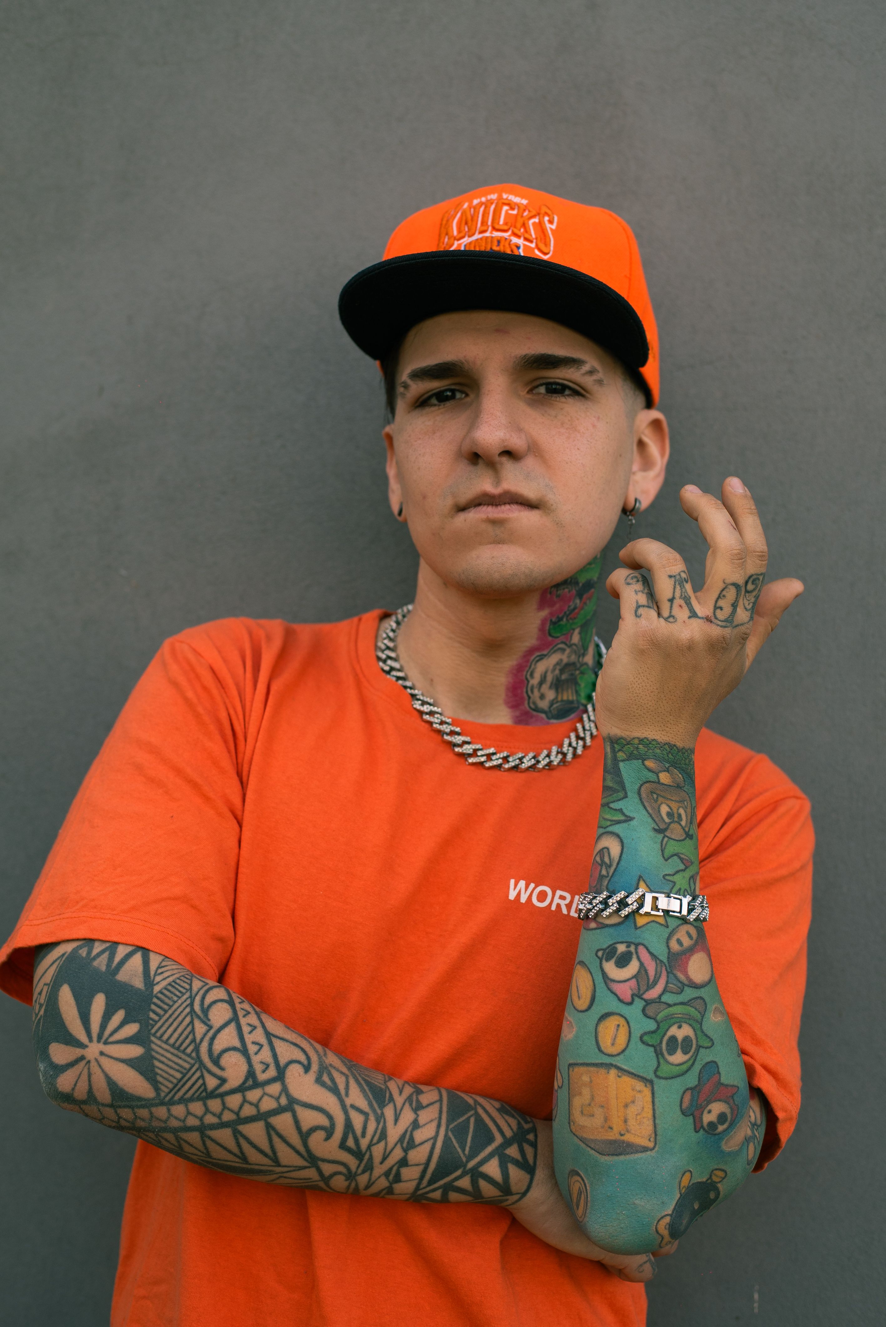 A young man in an orange shirt and cap shows off his colorful tattoos against a plain background.