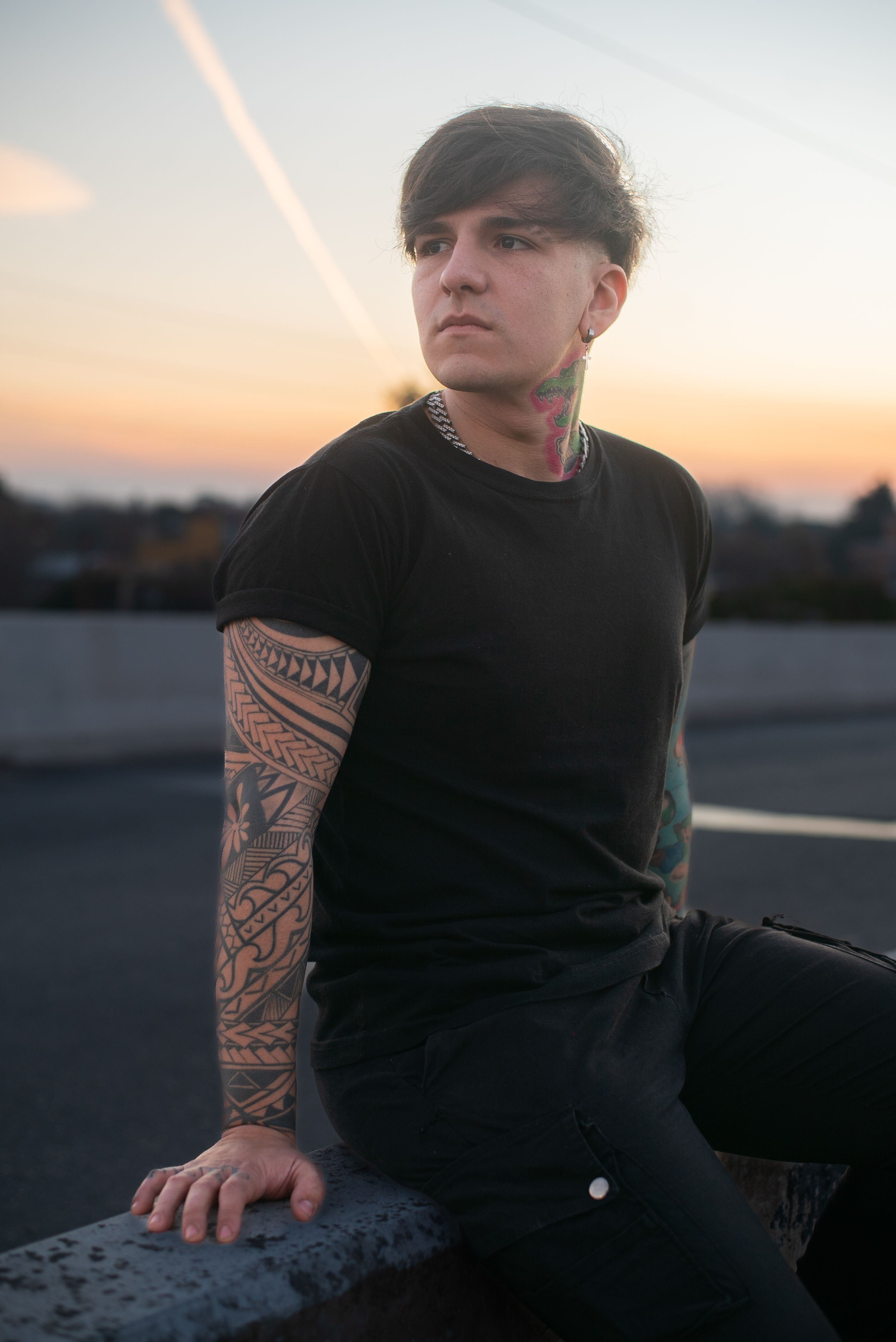 Stylish tattooed person sitting on ledge outdoors at sunset, exuding confidence and modern street style.