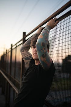 Tattooed person posing by a chain-link fence in an urban setting at twilight.