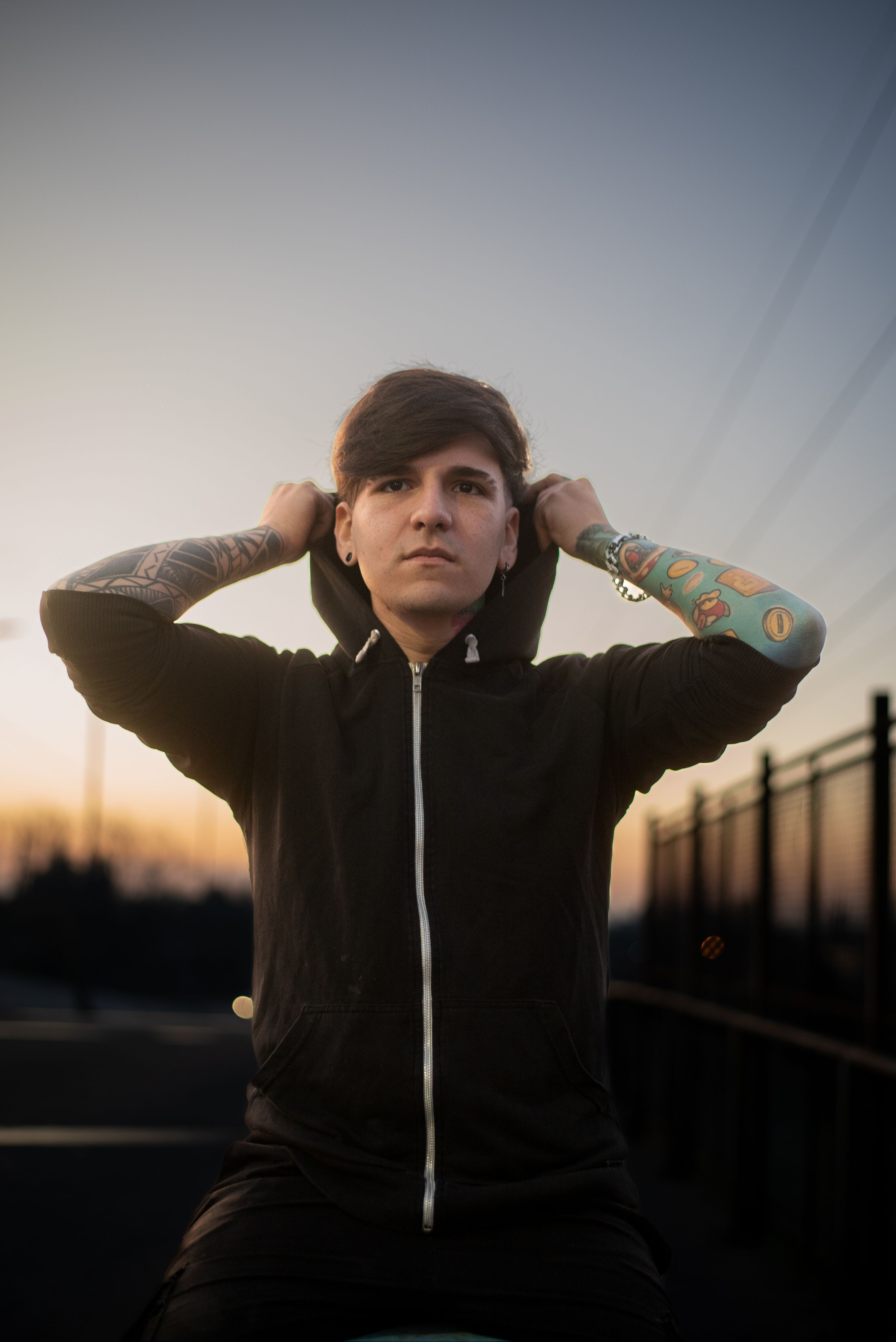 Portrait of a young man in a hoodie with tattoos, standing outdoors at sunset.
