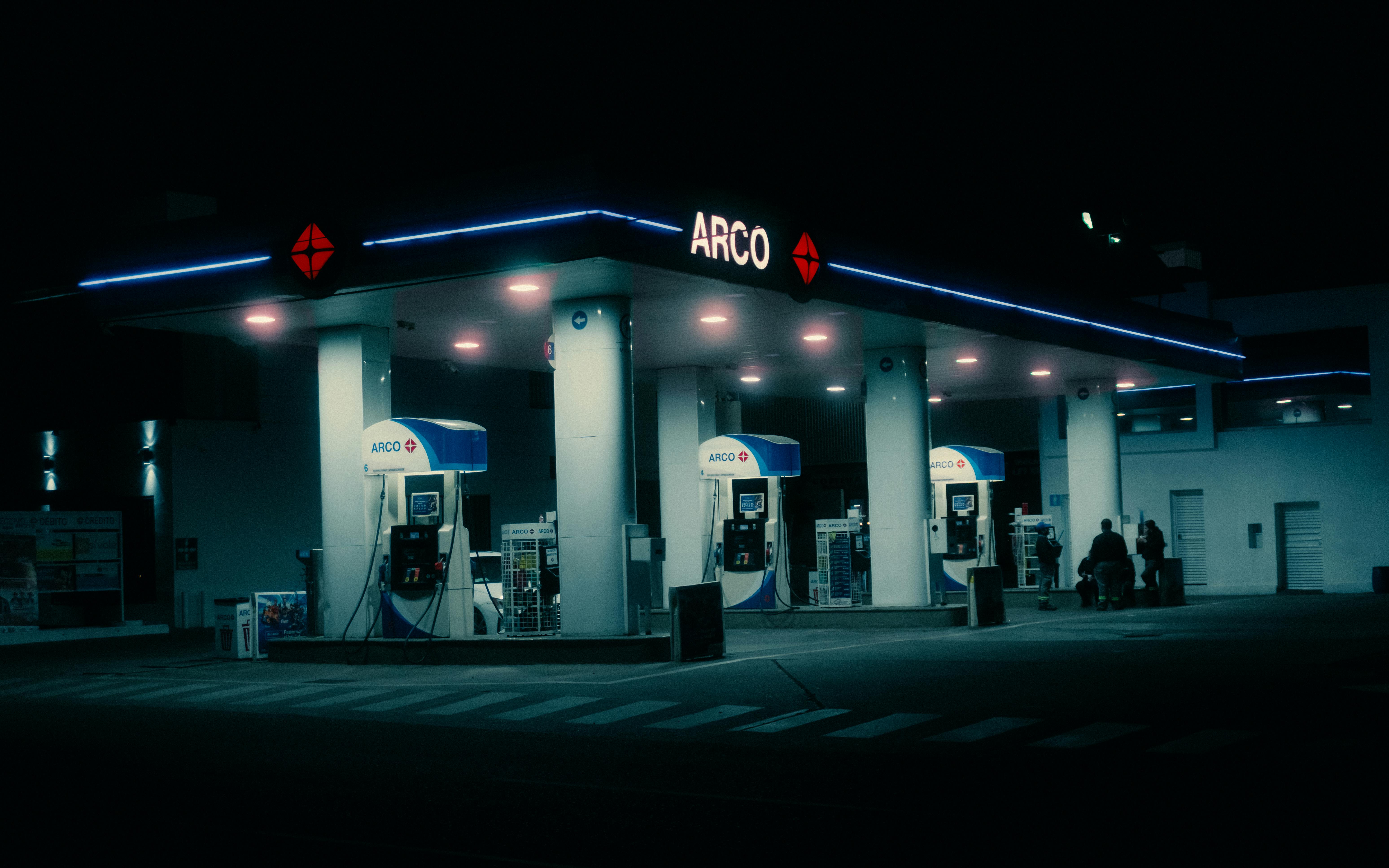 A cinematic night view of a modern ARCO gas station lit in neon against a dark backdrop.