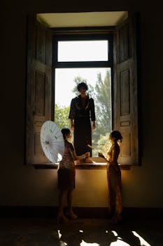 Three people in traditional attire standing by a sunlit window, creating artistic silhouettes.