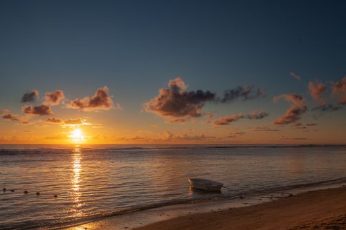Free A tranquil sunset scene with a row boat on a sandy shore, beautiful clouds, and calm ocean waves. Stock Photo
