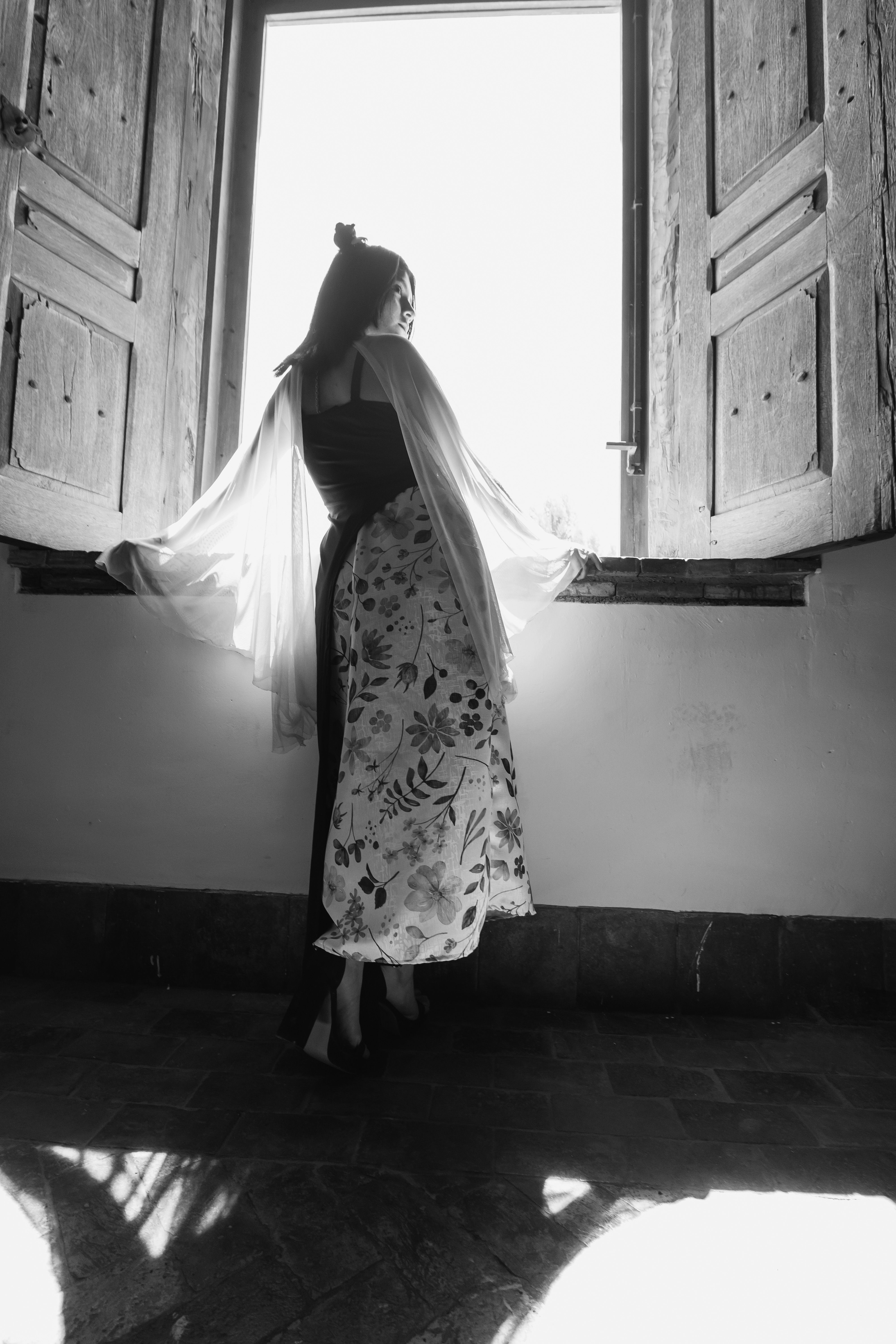 Black and white photo of an elegant woman in floral attire posing by a window indoors.