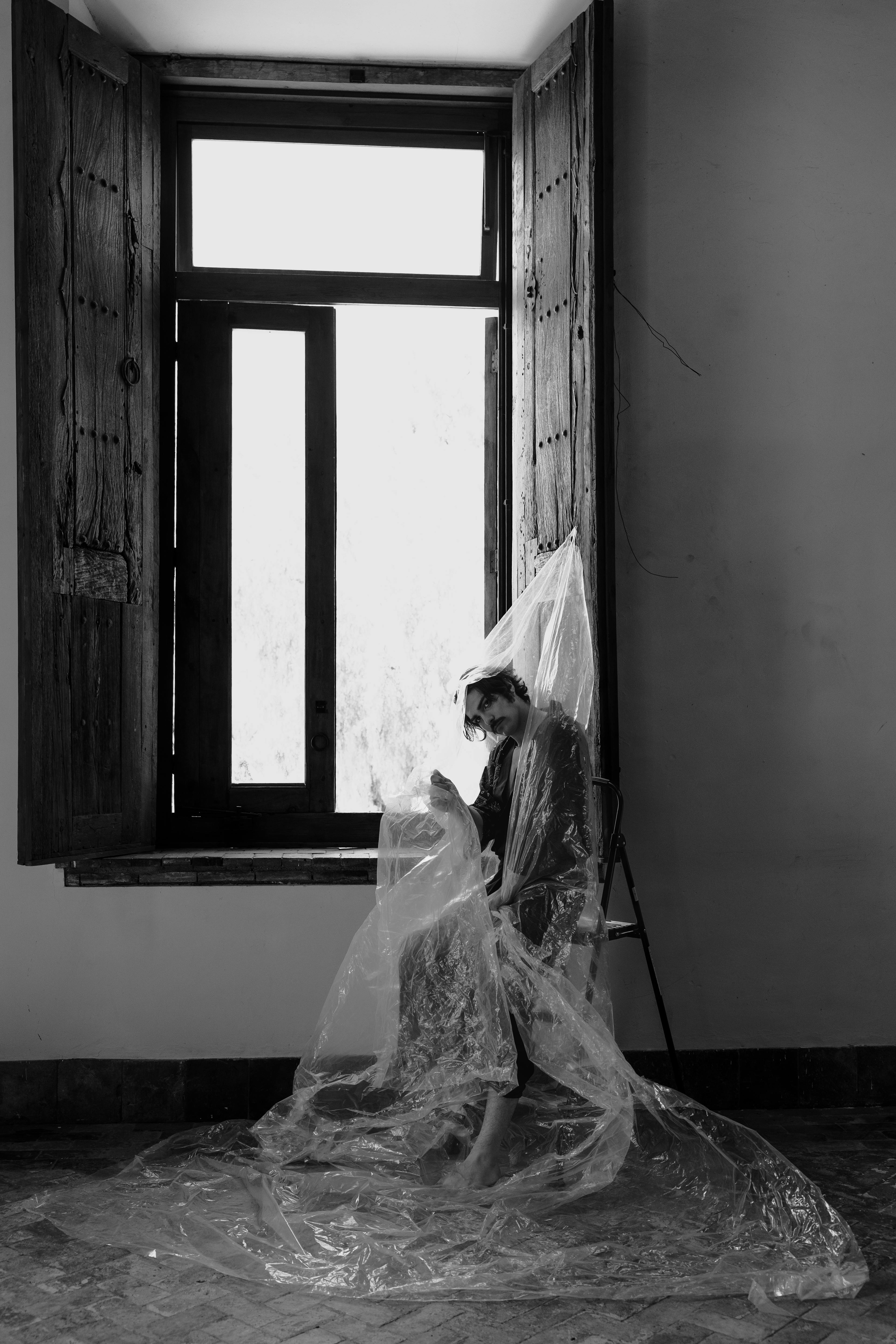 Free A dramatic black and white portrait of a person wrapped in plastic near an open window. Stock Photo