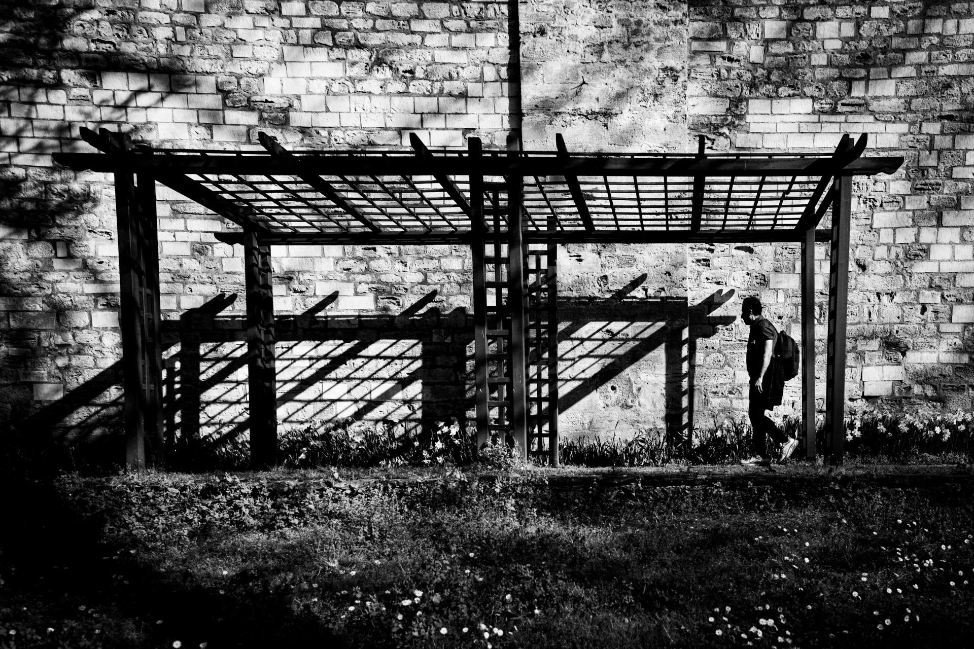 A person walks under a pergola casting geometric shadows against an ancient wall in İstanbul, Türkiye.