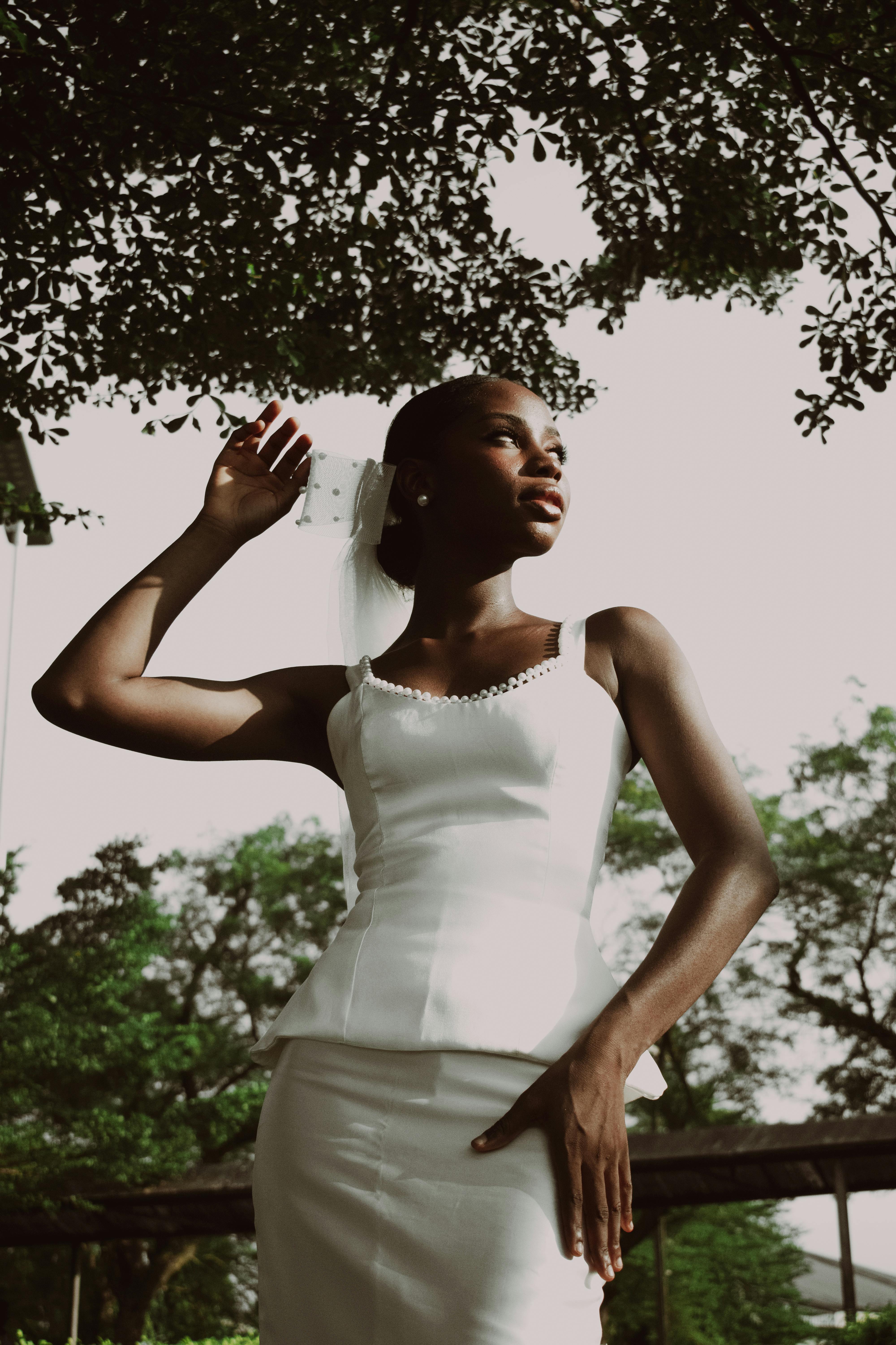 Elegant bride in a simple white dress posing outdoors under leafy trees.