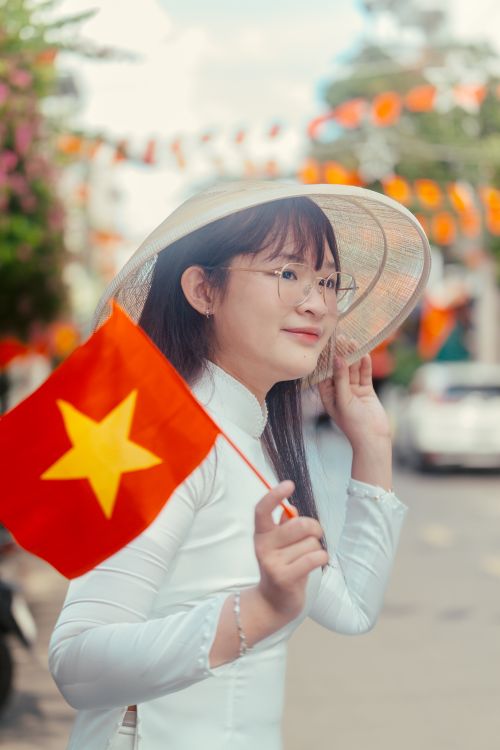 Free Young woman wearing traditional Vietnamese dress holding a Vietnam flag on a festive street. Stock Photo
