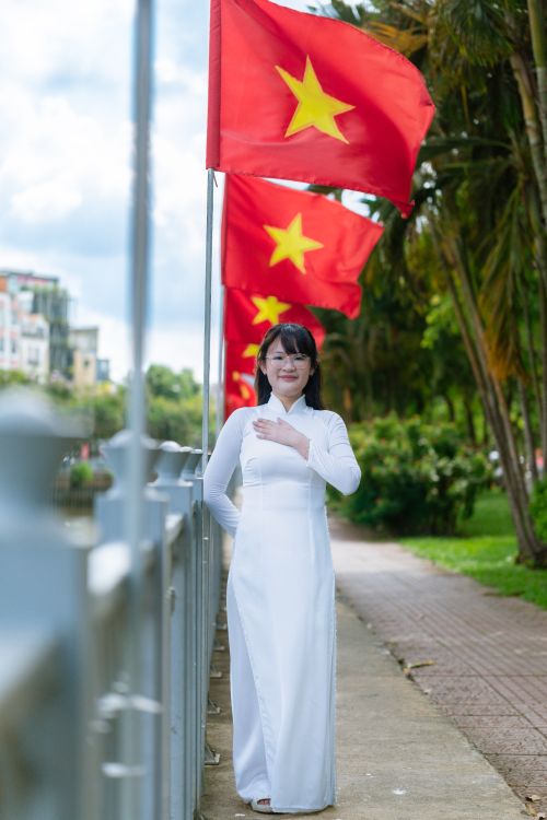 Free A Vietnamese woman in a white ao dai stands by red flags, symbolizing national pride. Stock Photo