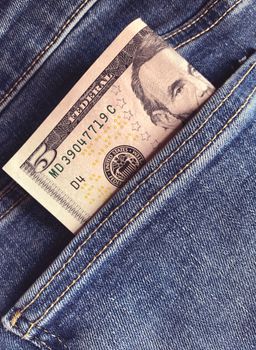 Close-up of a five dollar bill placed inside a blue denim pocket, symbolizing savings.
