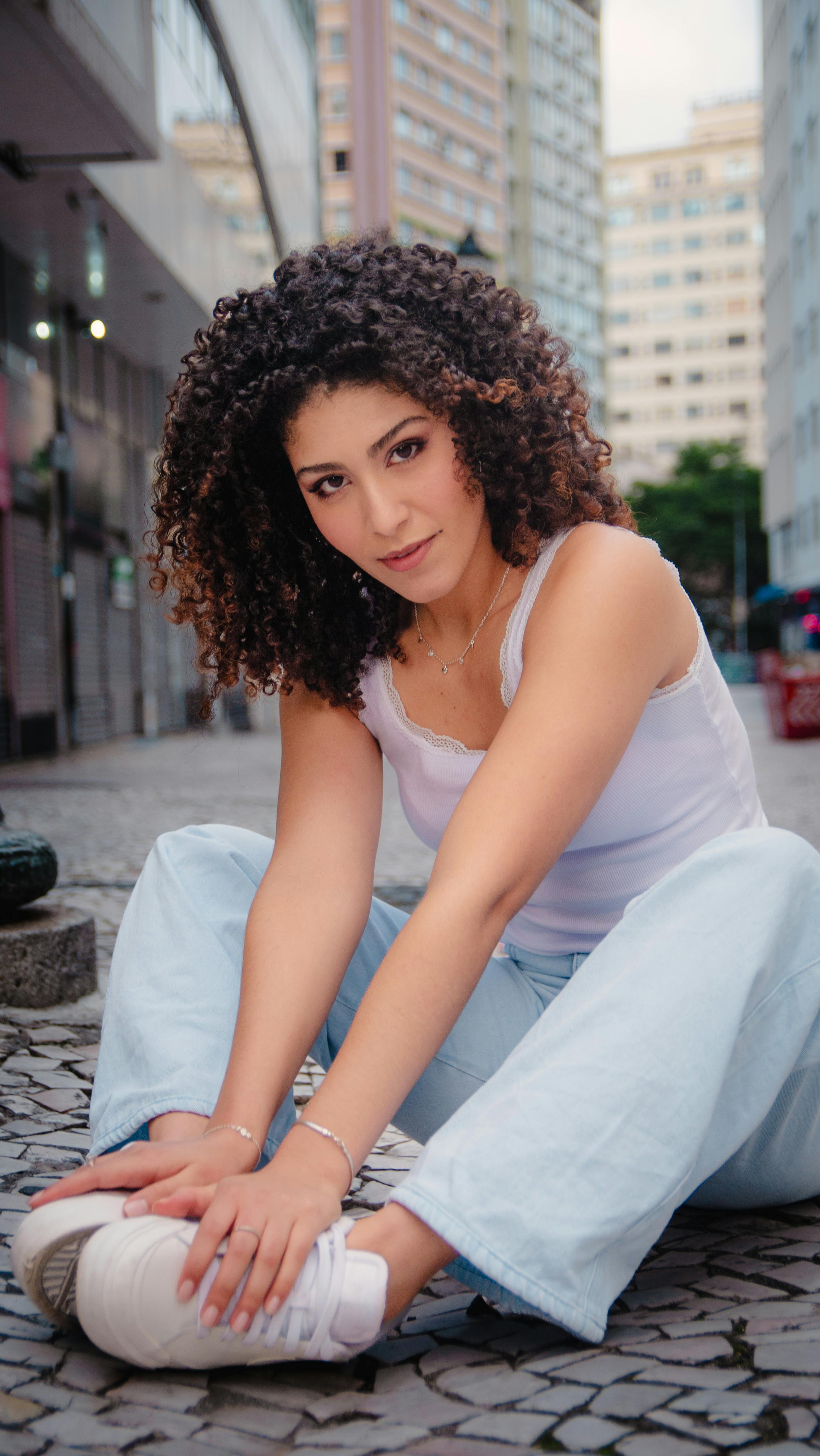 Young woman with curly hair sitting on a city street, wearing casual outfit.