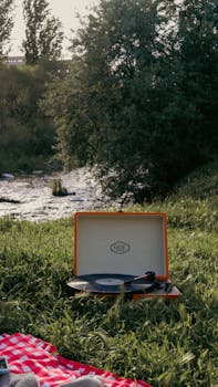 Retro record player set on a picnic blanket by a peaceful riverside, surrounded by lush greenery.