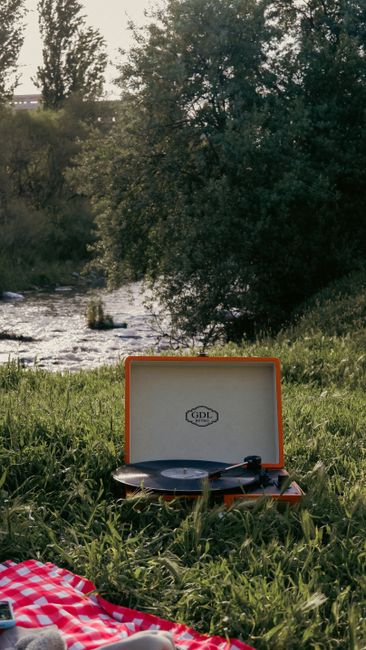 Retro record player set on a picnic blanket by a peaceful riverside, surrounded by lush greenery.