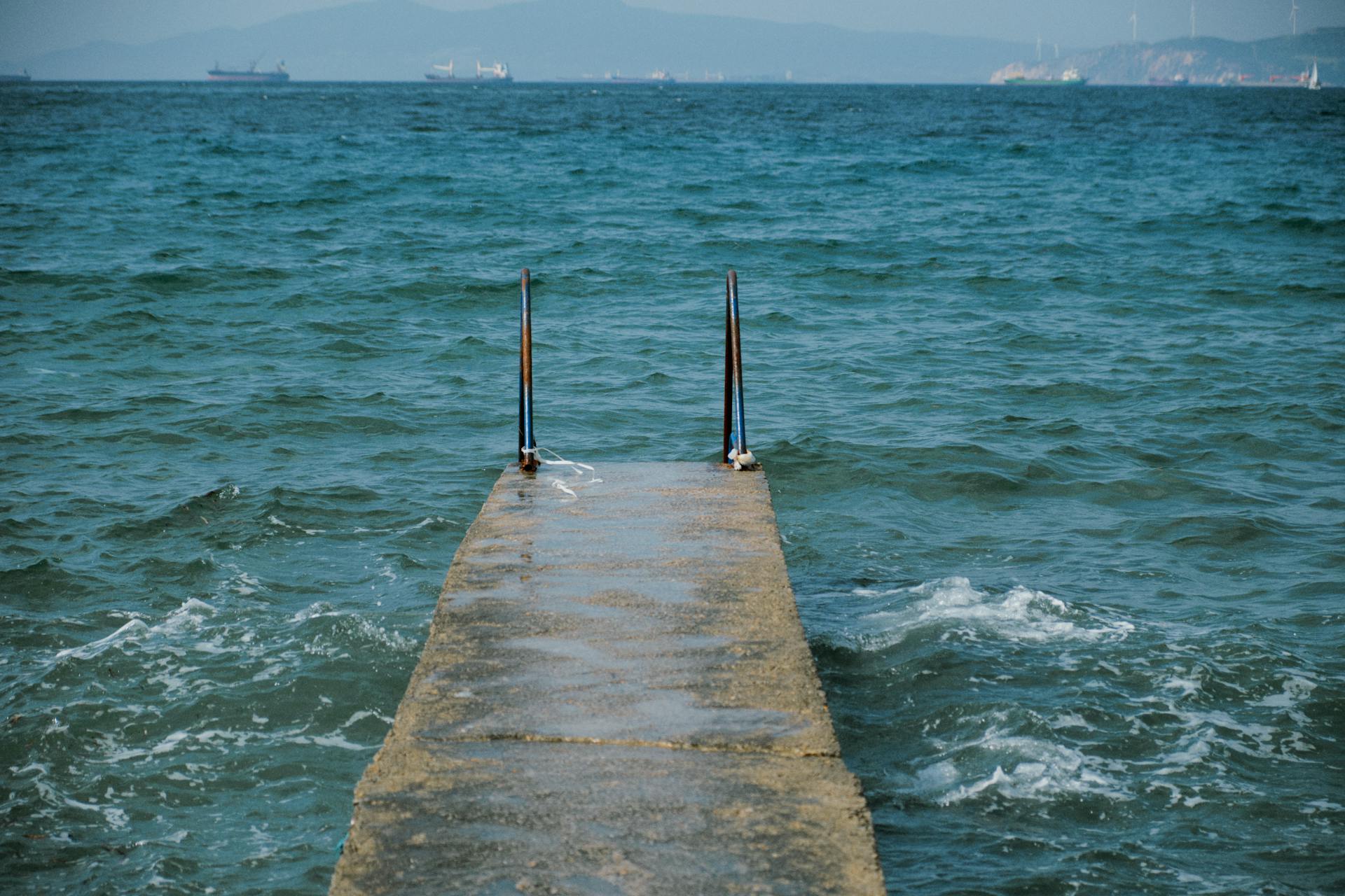 Serene jetty extending into the ocean with distant ships and mountains.