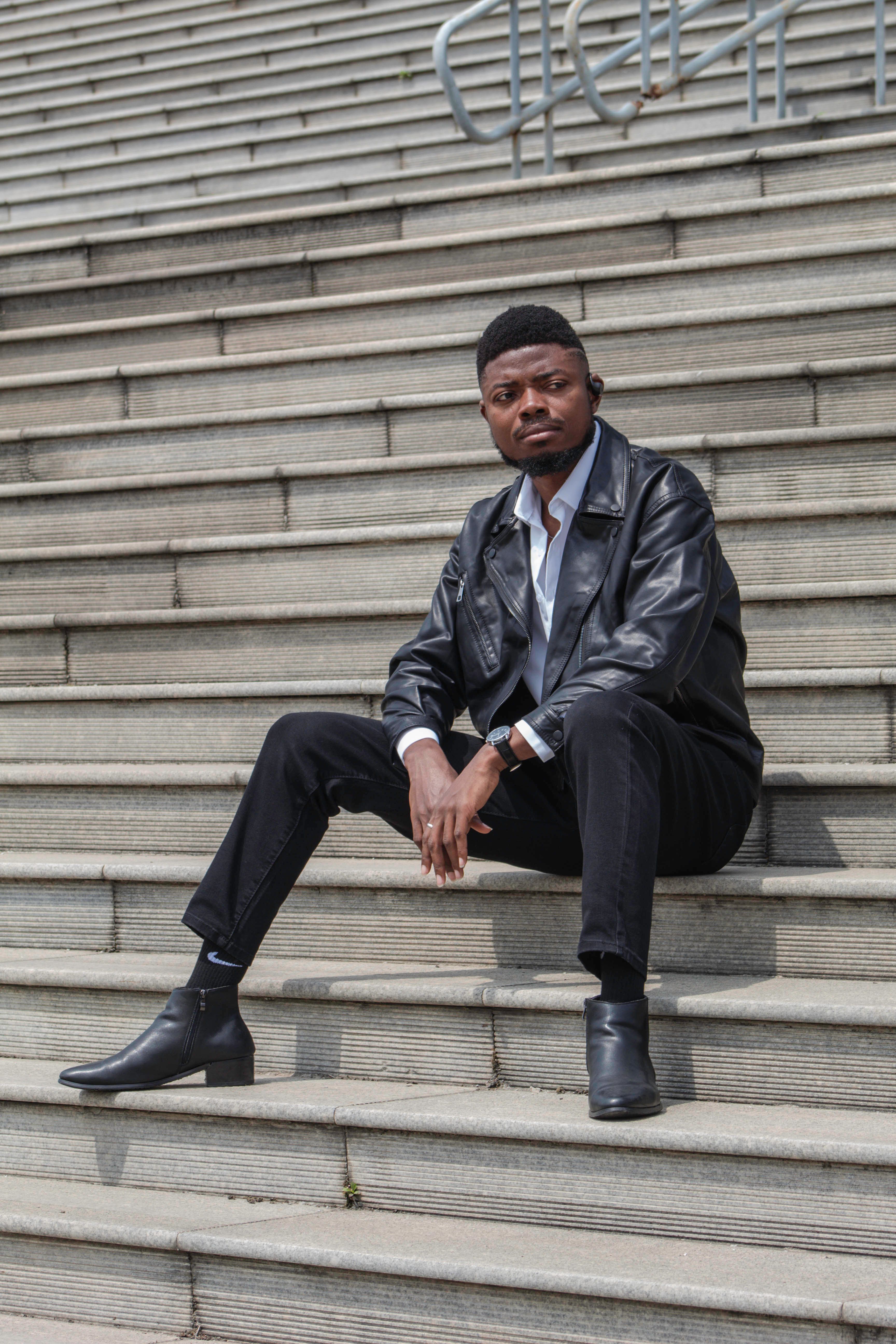 Fashionable young man in a leather jacket sitting on city steps, exuding confidence and style.