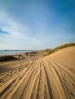 Wide sandy beach with tire tracks, blue sky, sea, and distant building; ideal summer coastal scene.