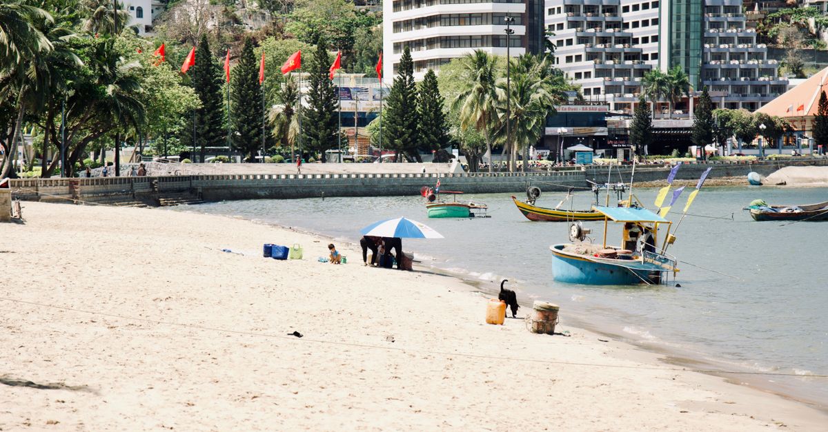 A vibrant beach scene with people enjoying the sand, boats docked, and a city skyline in the background.