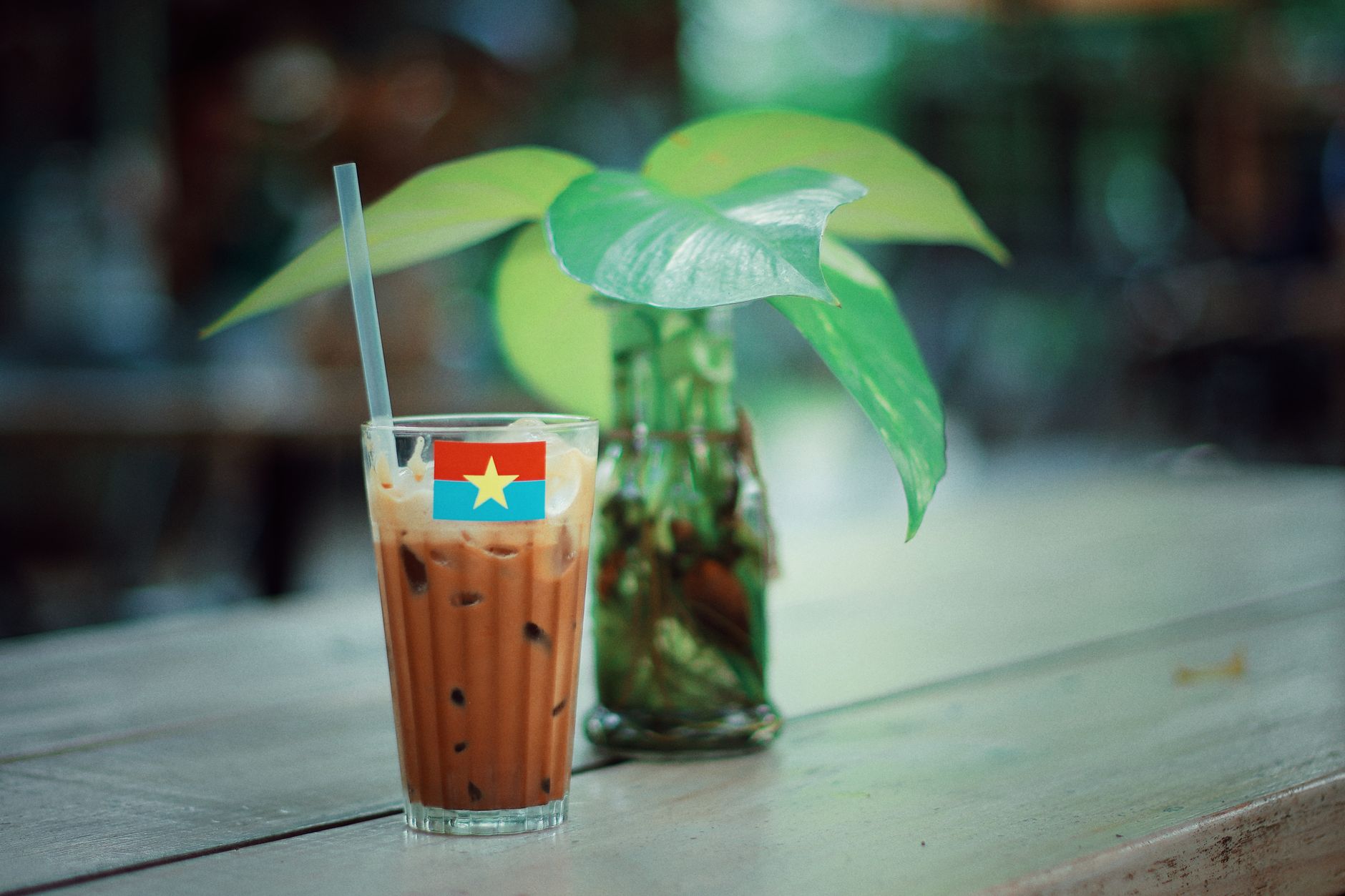 Glass of iced coffee with straw on wooden table next to greenery in a cafe setting.