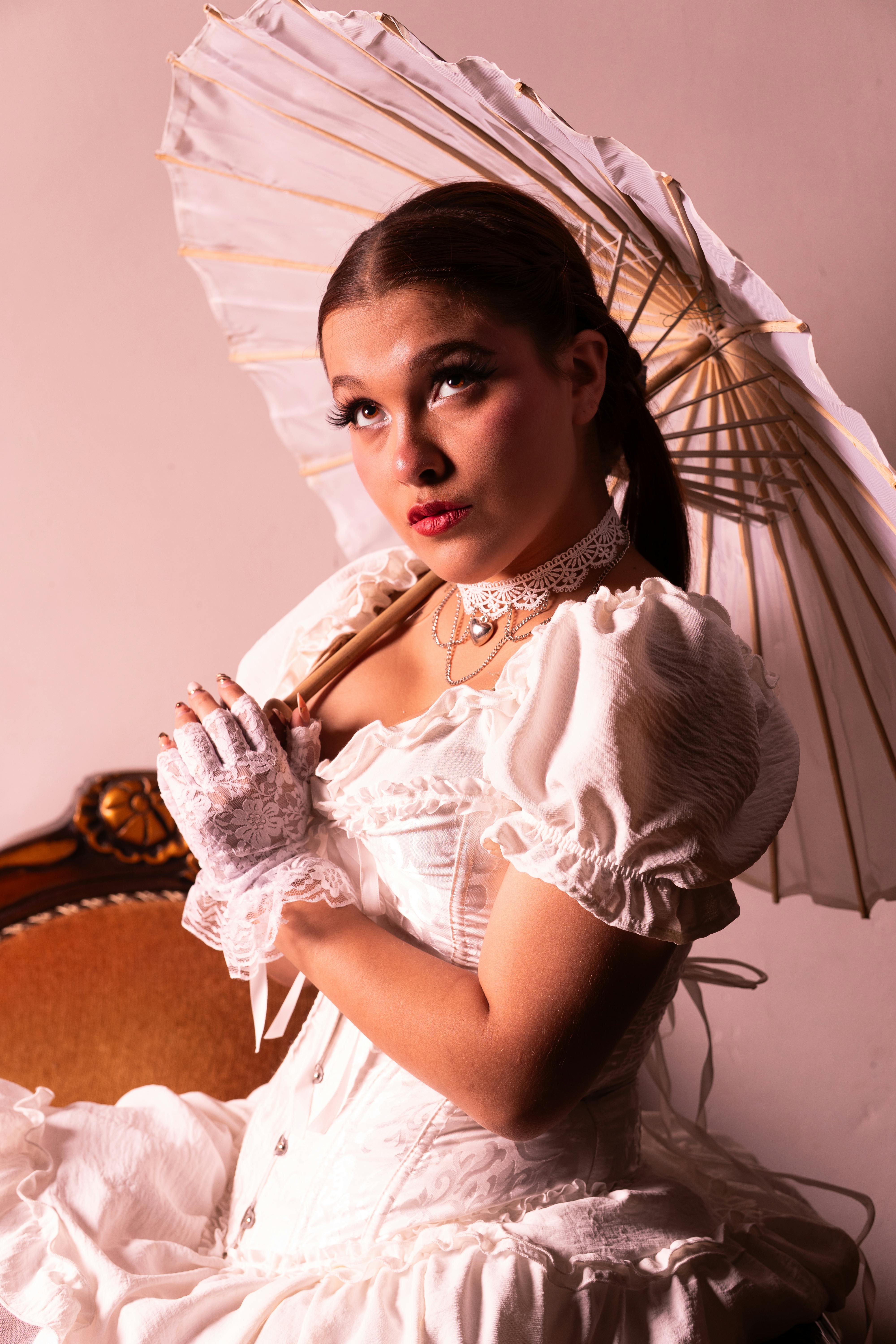 Bezpłatne Inspirowany stylem vintage portret kobiety w białej sukni i z parasolką, przywodzący na myśl klasyczną elegancję. Zdjęcie z galerii