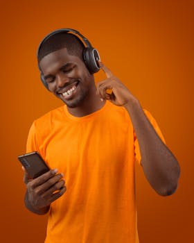 Smiling man in orange shirt listening to music on headphones with smartphone.