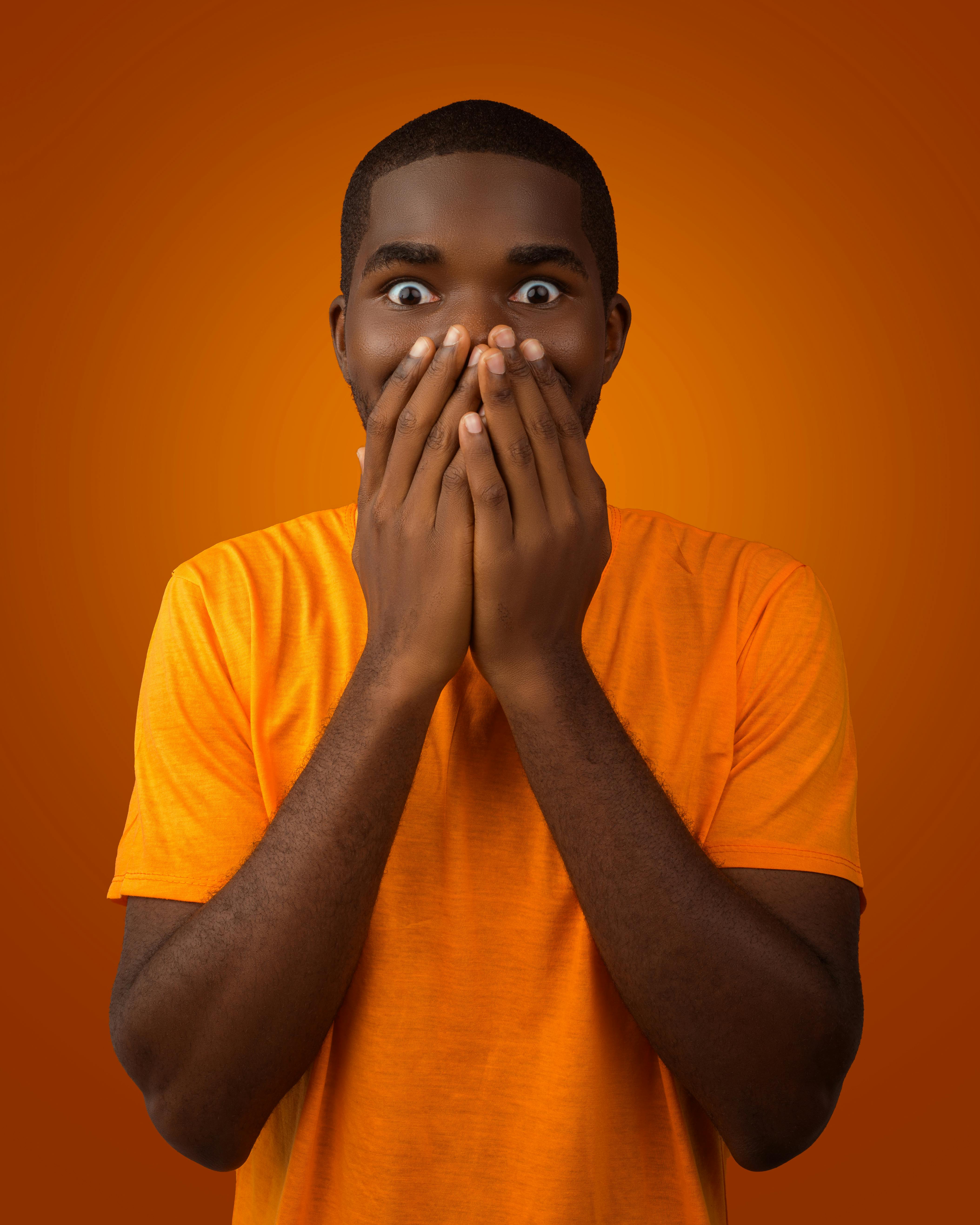 A surprised man with wide eyes covering his mouth in shock against an orange background.