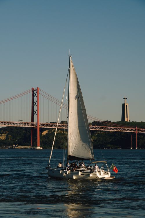 Free A sailboat glides past the 25 de Abril Bridge in Lisbon, Portugal, as the sun sets. Stock Photo