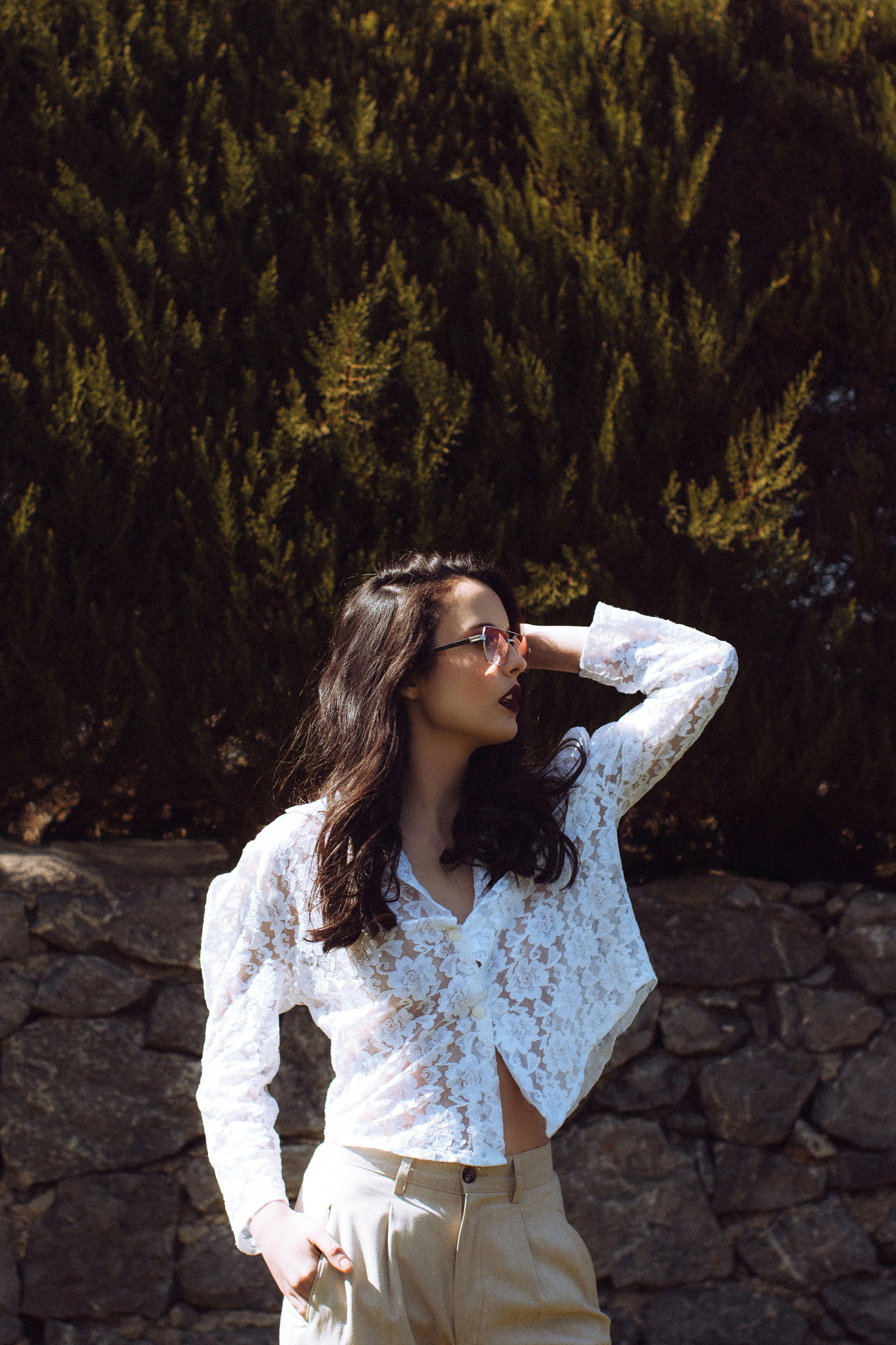 Fashionable woman in a white lace blouse posing confidently outdoors with greenery background.