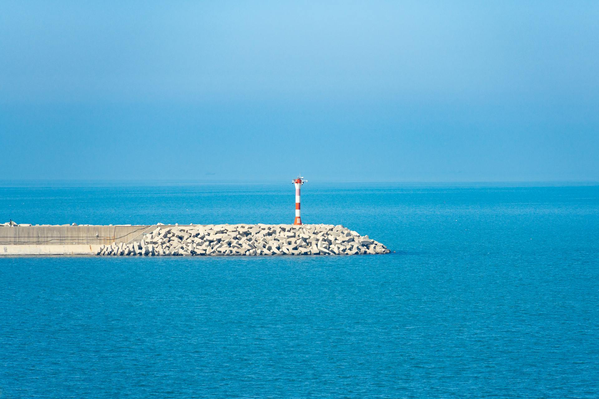 A lighthouse on a rocky breakwater under a bright blue sky and calm sea, ideal for maritime themes.