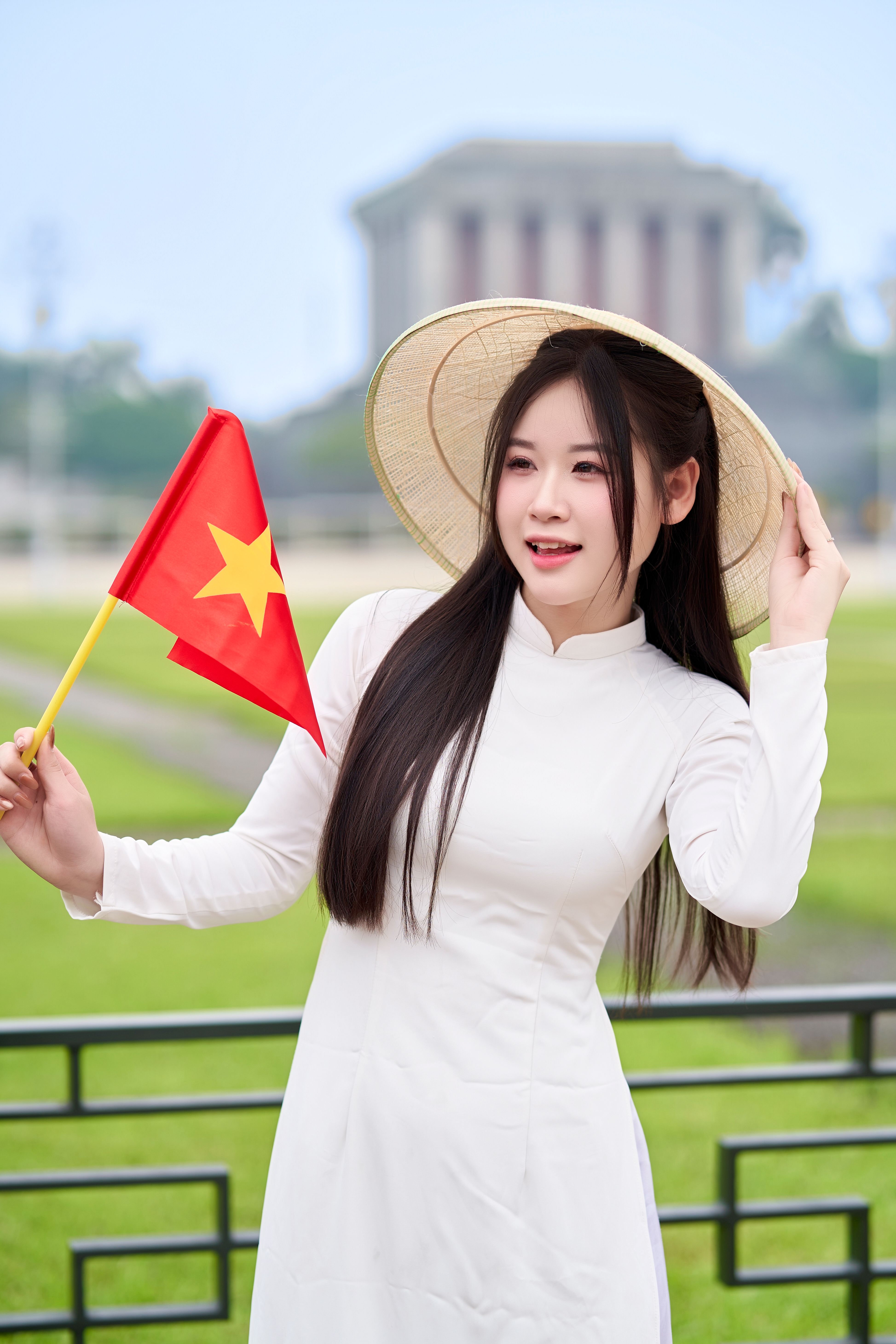 Woman in white áo dài with Vietnamese flag outdoors near historic landmark.
