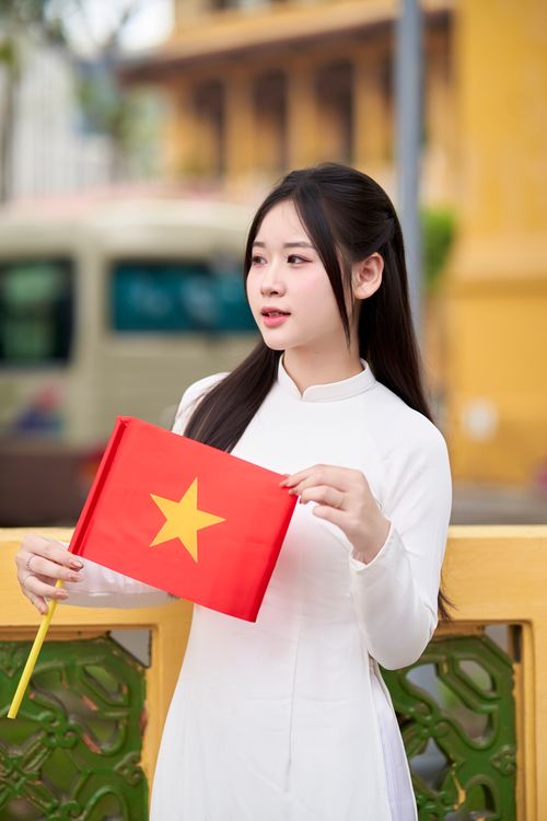 Free A young woman in a white Ao Dai holds the Vietnamese flag outdoors, symbolizing national pride. Stock Photo