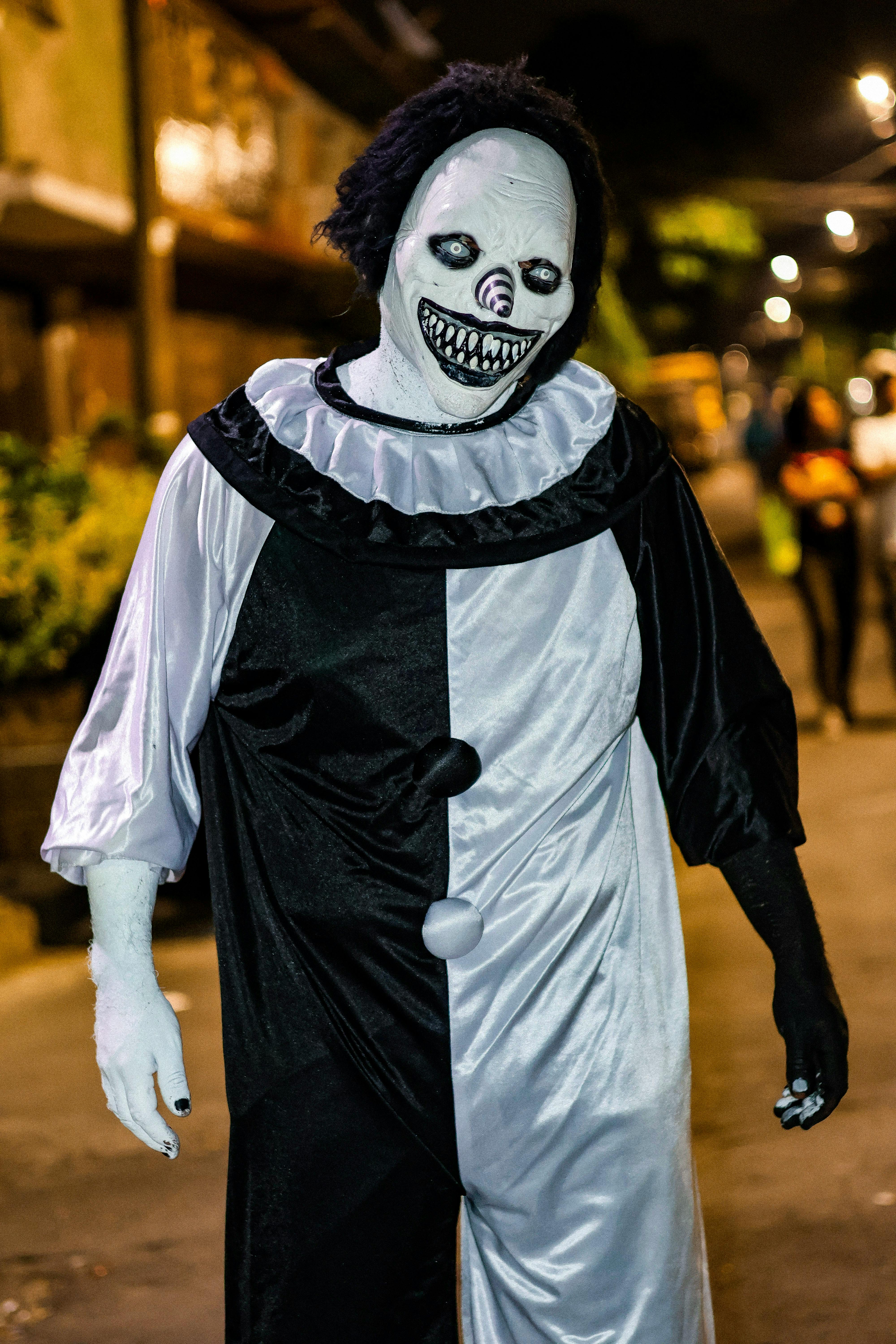 A person in a frightening clown costume walks through a dimly lit street at night.