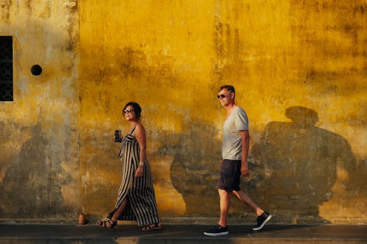 A couple walking in casual wear past a vibrant yellow wall, casting shadows, on a sunny day.