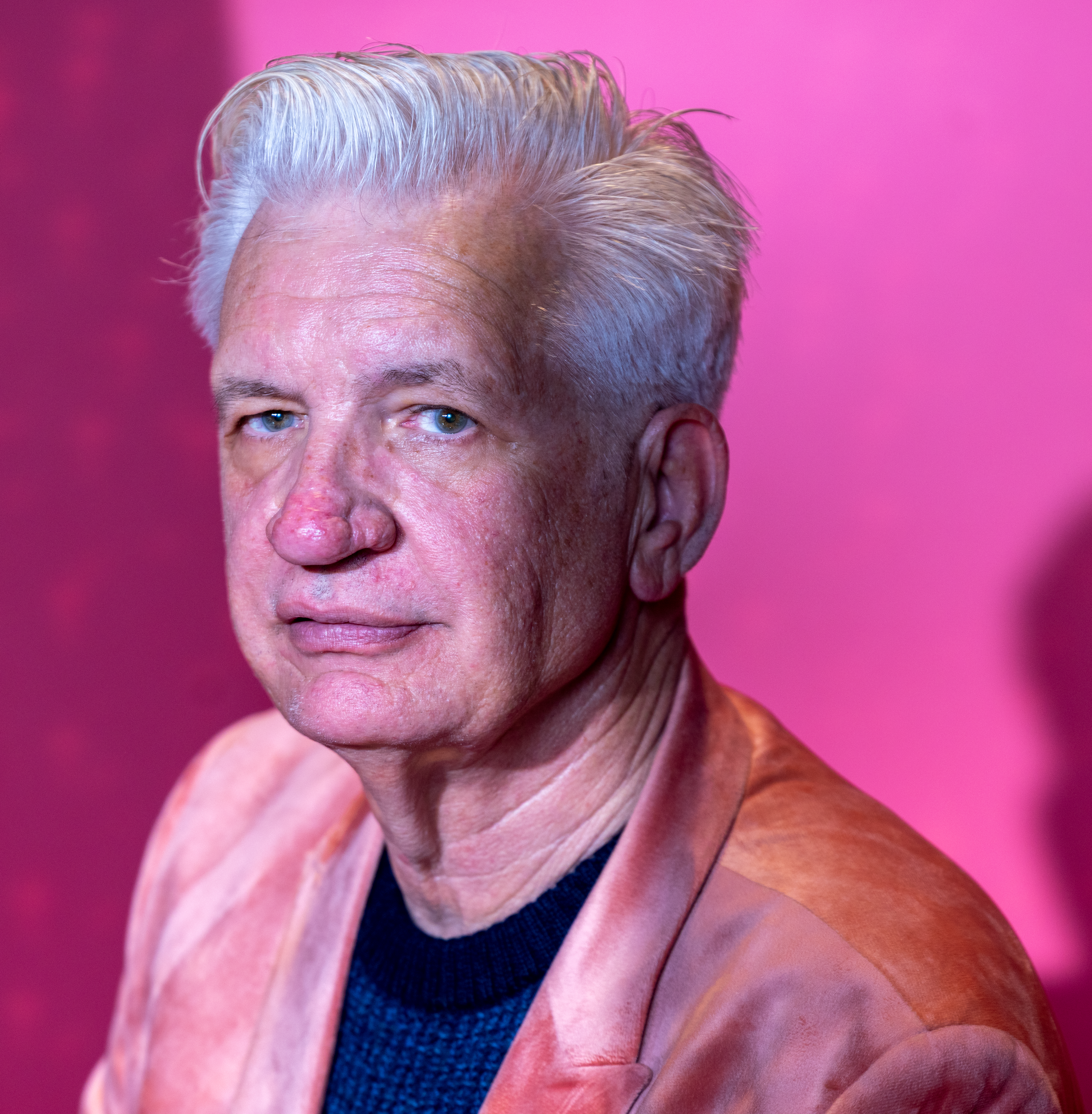 Elderly man with white hair and pink jacket against pink backdrop.
