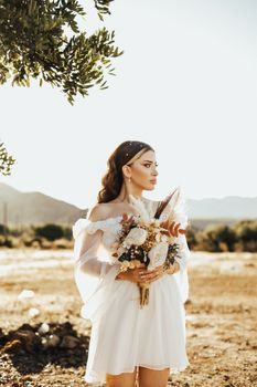 A bride in a white dress holding a floral bouquet in a sunny, natural landscape.