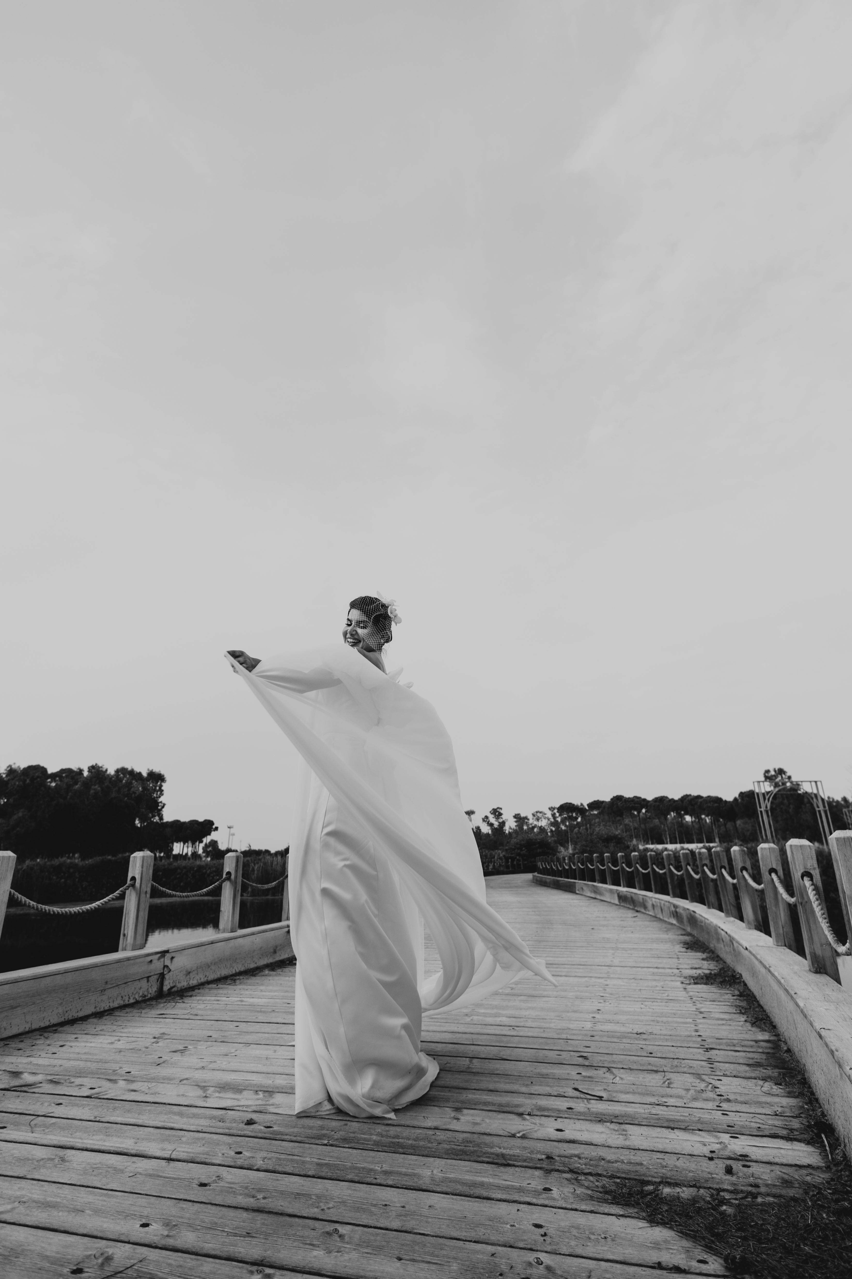 Elegant figure in flowing dress captured on a serene wooden bridge in black and white.