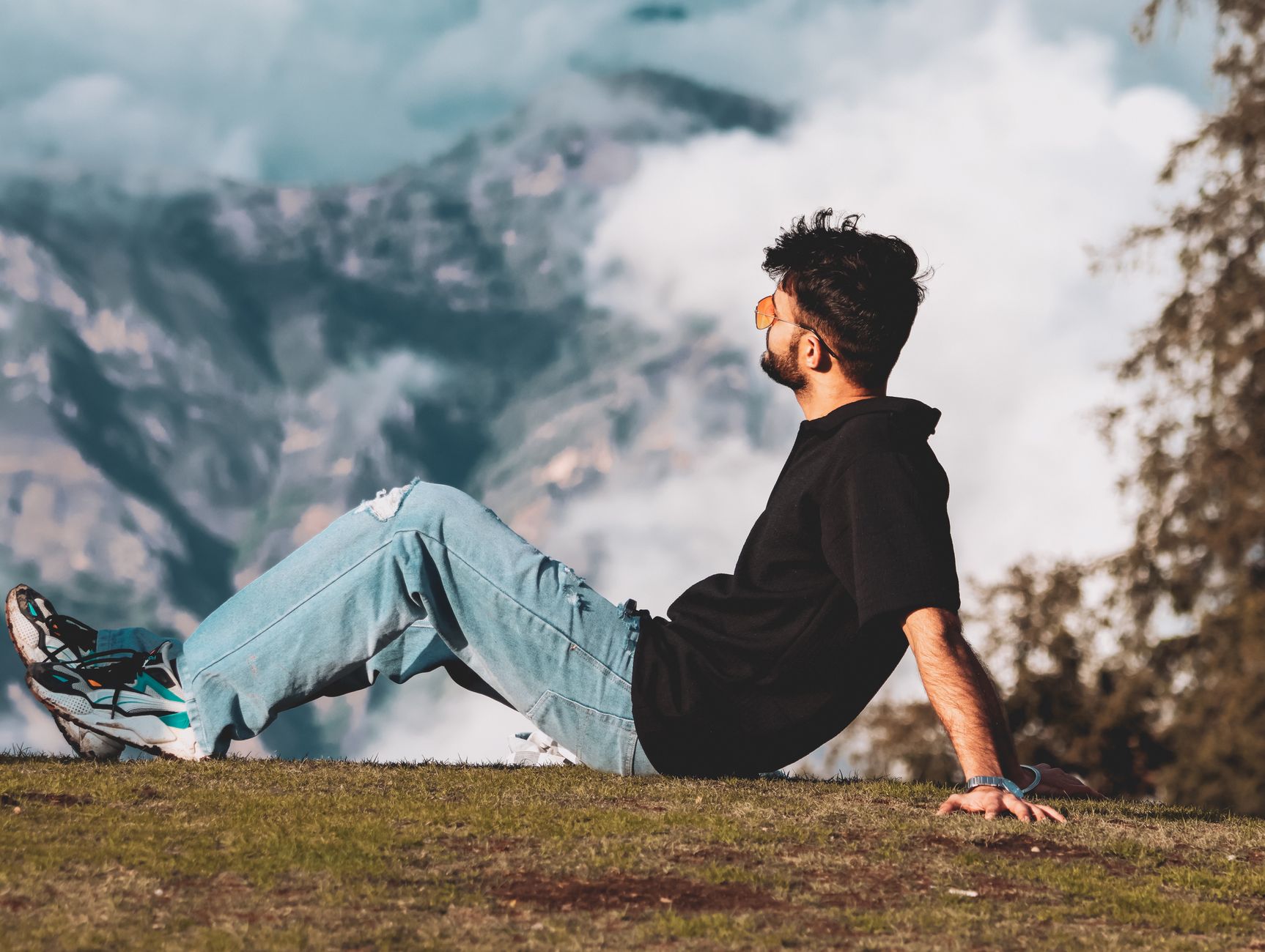 A young man relaxes on a hillside, enjoying scenic Himalayan mountain views with clouds.