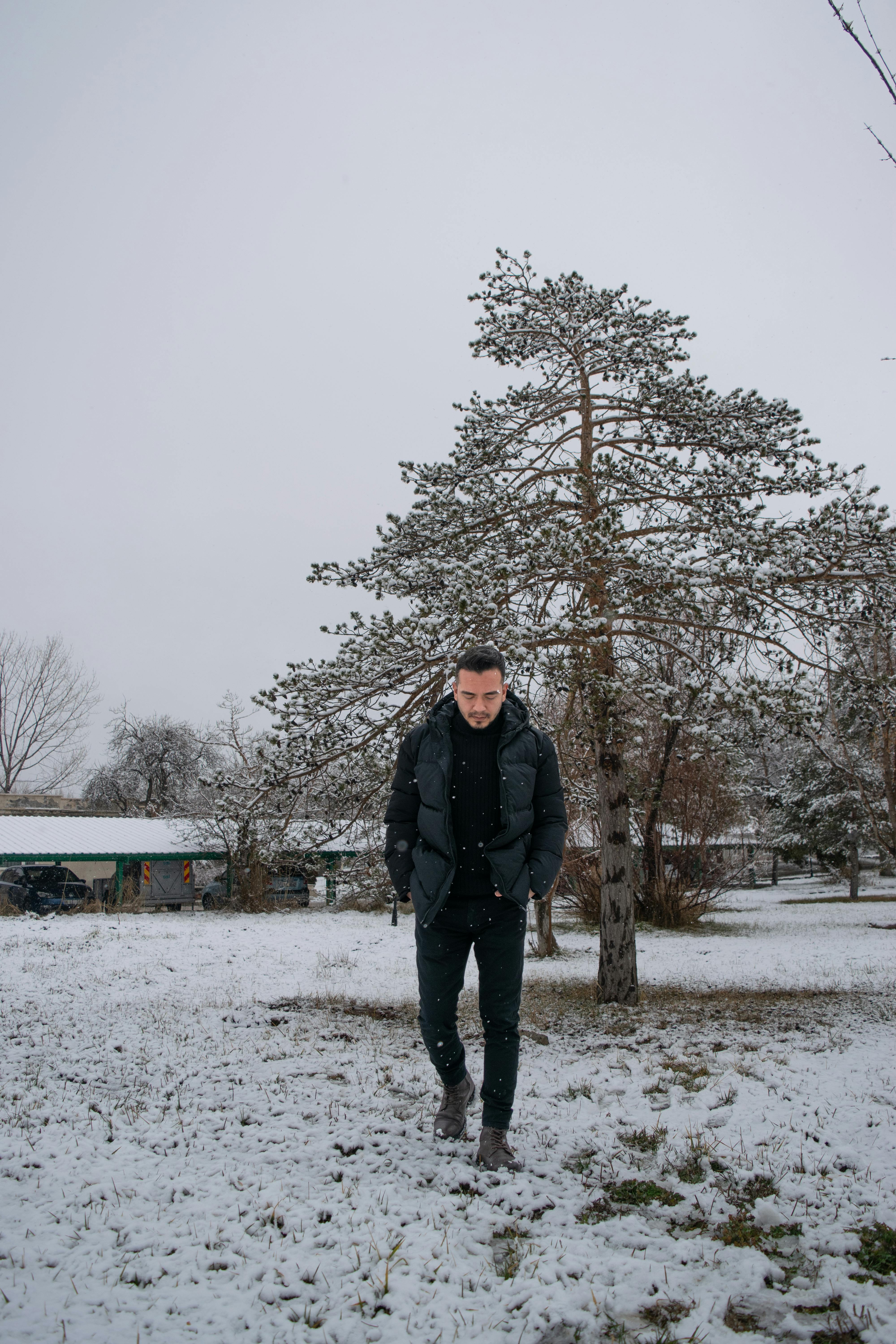 A man in winter clothing walks through a snowy park with trees in the background.