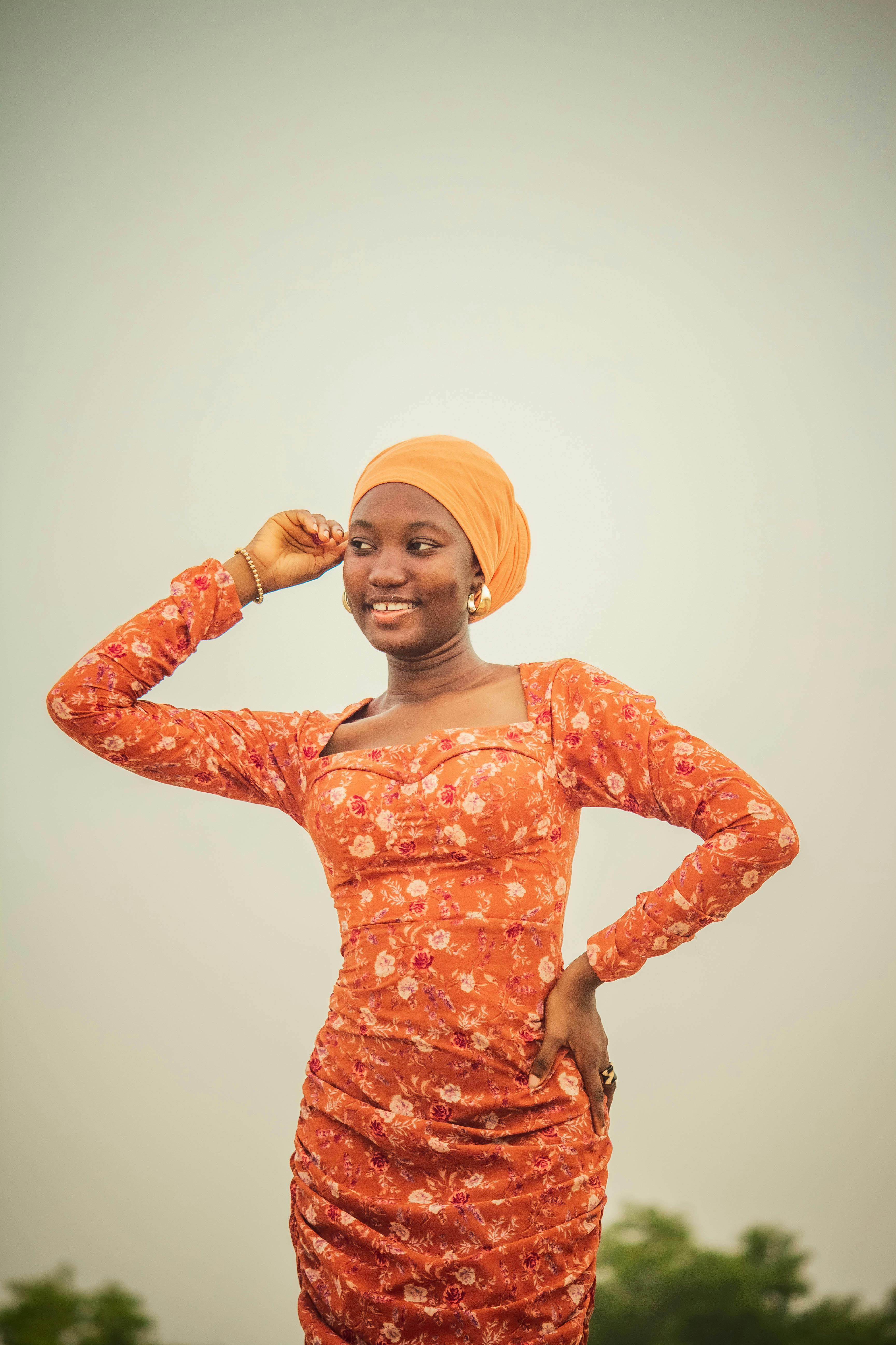 A woman stands confidently in an orange floral dress, smiling joyfully against the sky.
