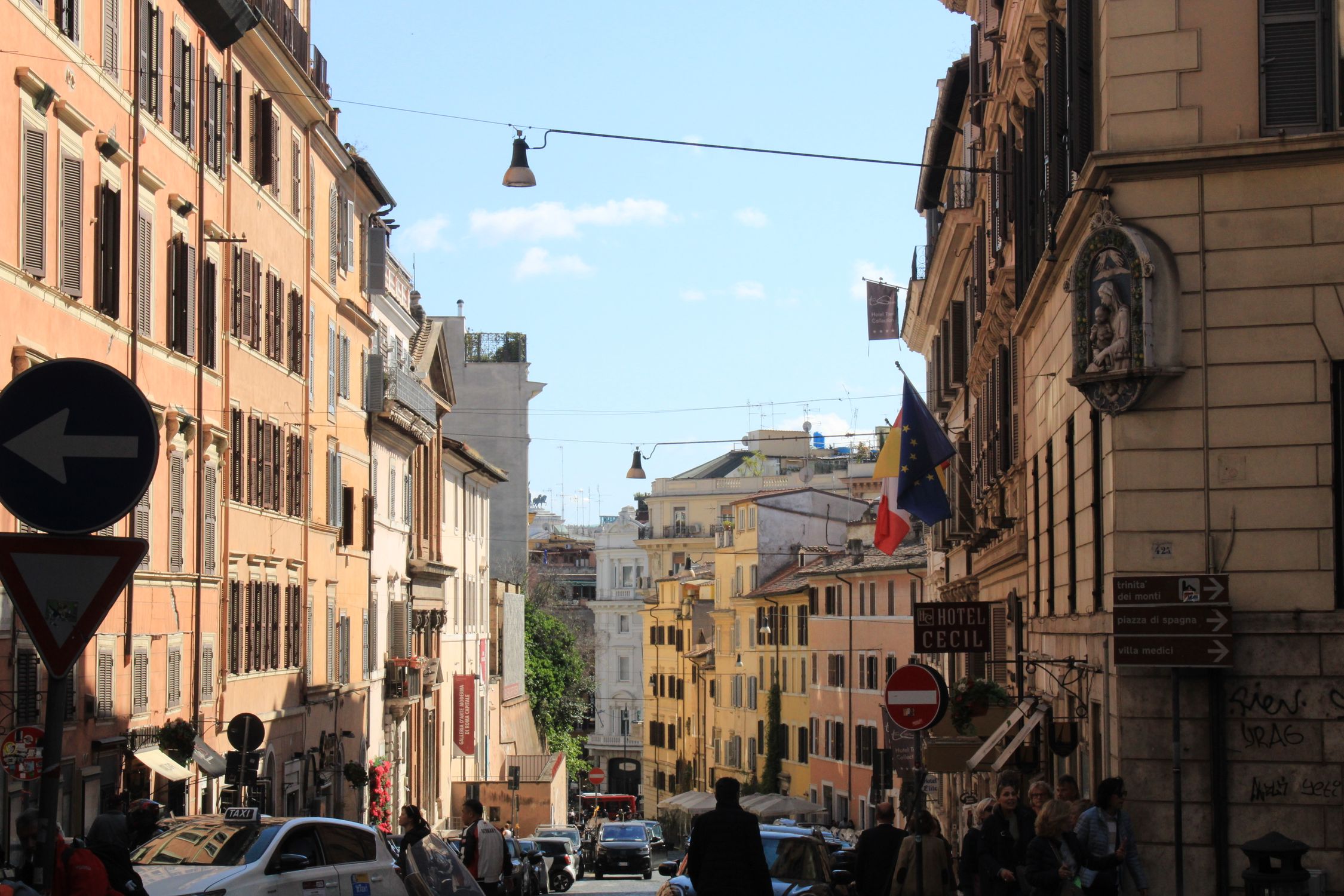 A bustling street in Rome with historic buildings, flags, and a lively atmosphere under a clear sky.