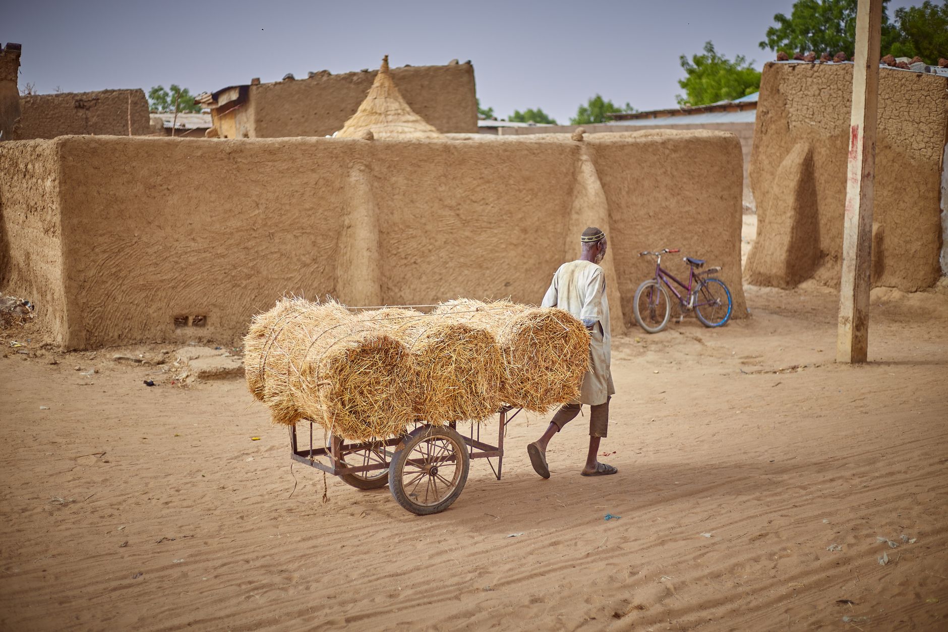 A man pulls a cart full of hay through an African village with adobe buildings.