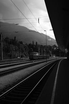Black and white image of a railway station with mountains in the background.