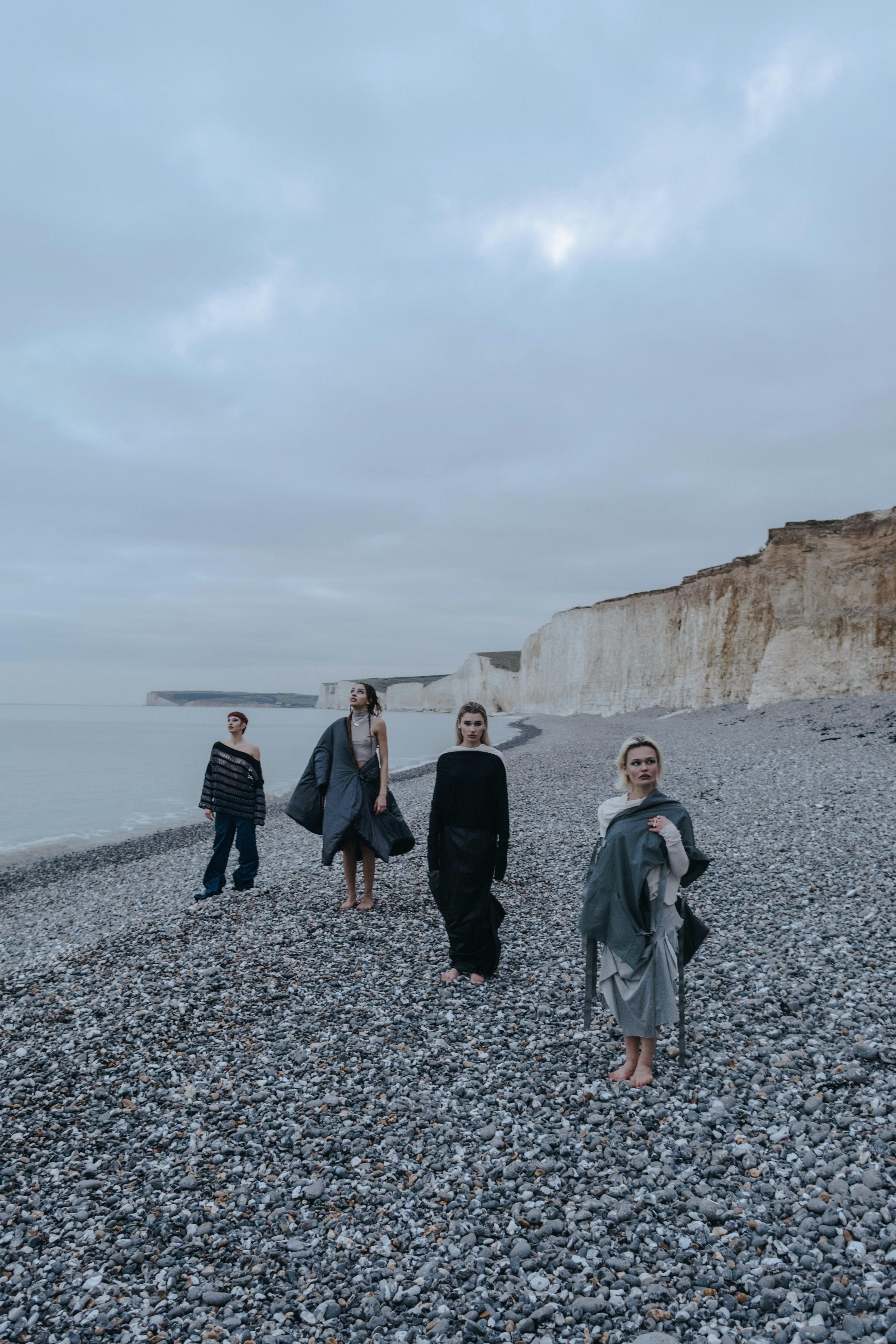 Group of adults in stylish attire walking on a pebble beach by white cliffs under a cloudy sky.