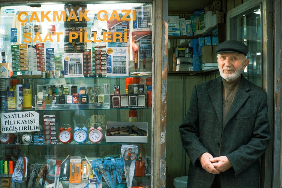 An elderly shopkeeper stands beside a traditional store in Istanbul selling batteries and lighters.