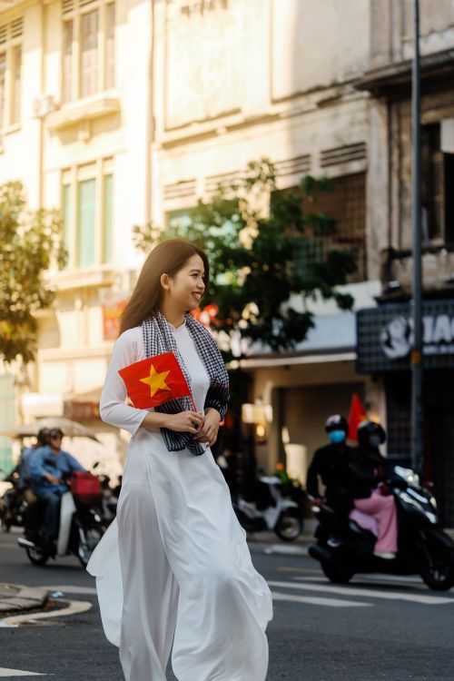 Free Young woman in white dress holding Vietnamese flag, walking on a bustling city street. Stock Photo