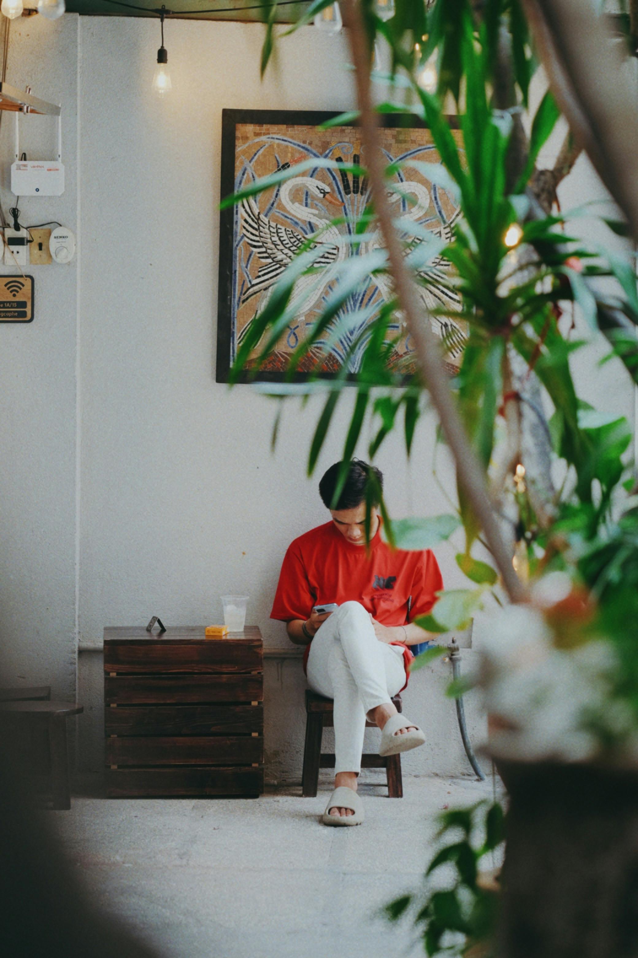 Gratuit Persoană așezată într-o cafenea confortabilă, înconjurată de verdeață și artă, oferind o atmosferă liniștită. Fotografie de stoc